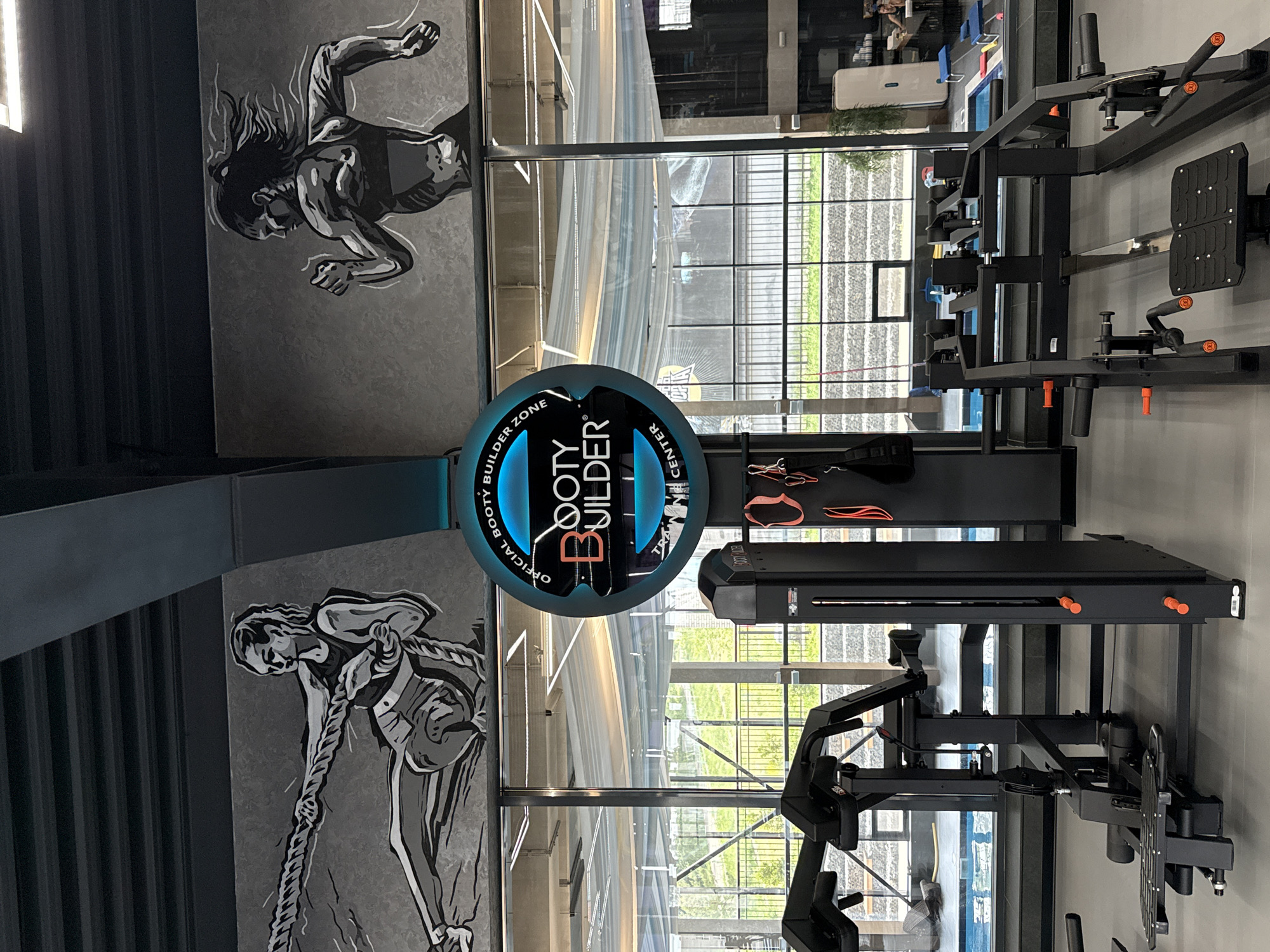 The image depicts an indoor gym space. The walls feature grayscale graphic art depicting individuals engaged in physical activity. A prominent sign reading "Booty Builder Center" is centrally positioned. The space is equipped with various pieces of gym equipment, including a leg press machine. Natural light enters through large windows, revealing an exterior view of a city street with greenery and architecture. The gym is modern, clean, and well-lit. Although I see the name of the city "Votkinsk" in your prompt, this information is not visibly evident in the image and is therefore not reflected in the description.