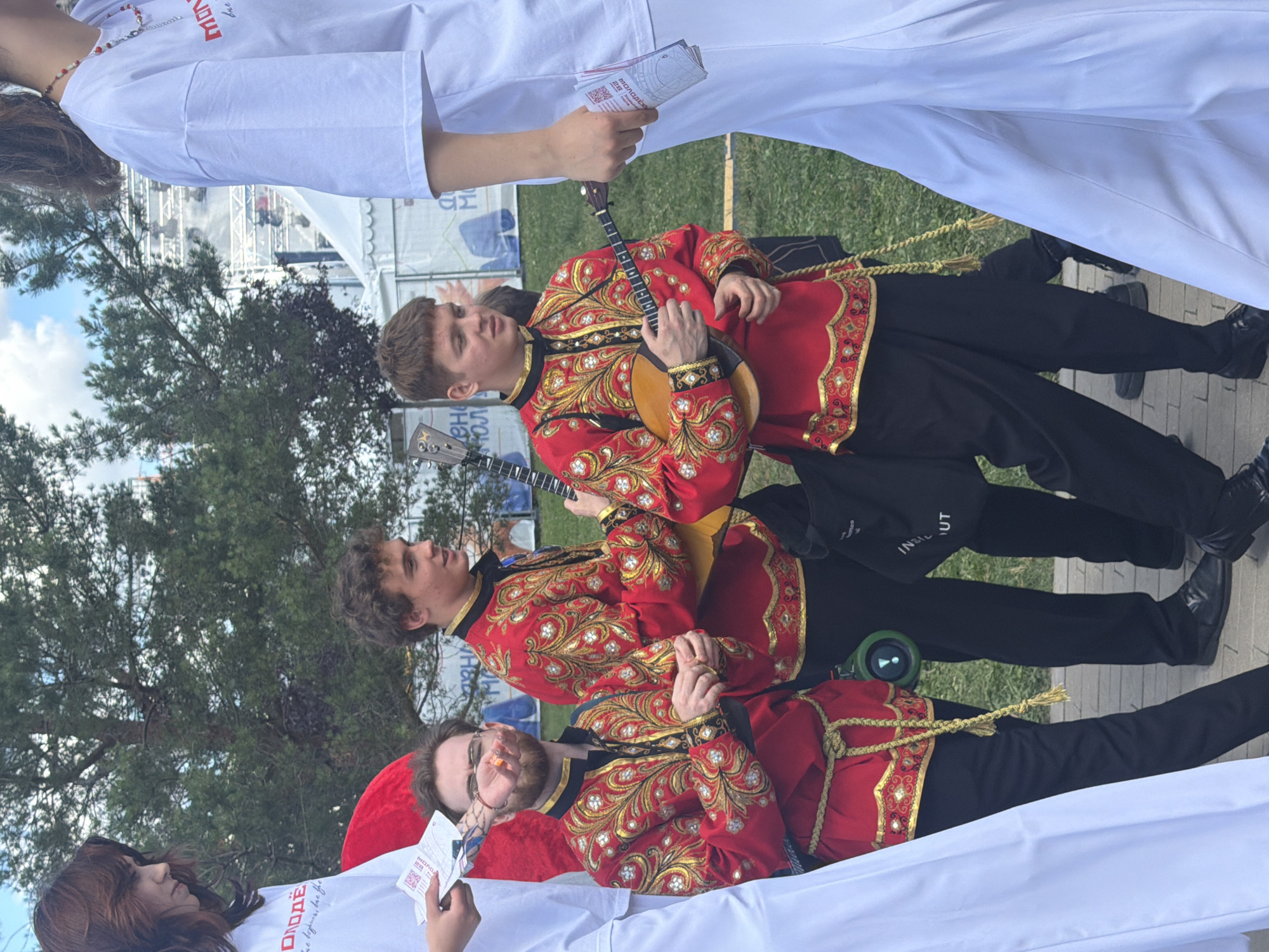The image shows an outdoor scene with several people dressed in traditional Russian attire. In the foreground, three young men are wearing red tunics with gold embroidery and black pants. Two of them are holding balalaikas, a type of Russian stringed instrument. A young woman in a white garment is to the left, holding papers. Another person in white, possibly a woman, is on the far right, also holding papers. The background suggests a public event with tents and stage equipment. The ground is paved with bricks, and there is green grass and trees visible. The sky is partly cloudy.