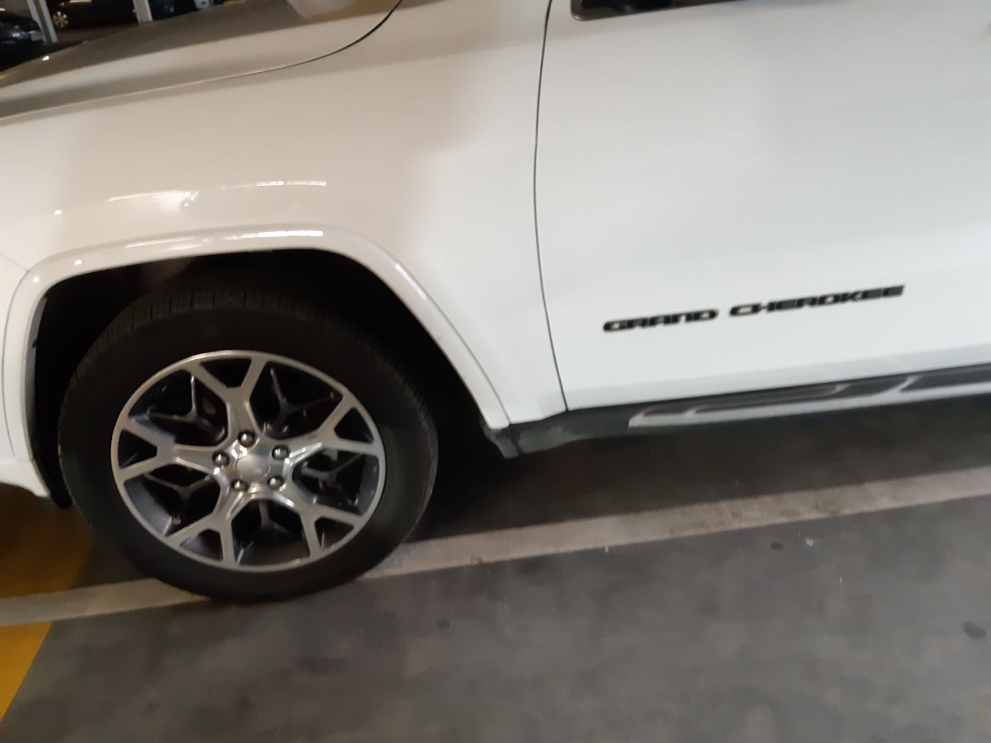 A close-up shot shows the front passenger side of a white Jeep Grand Cherokee. The car is parked indoors, possibly in an underground parking garage, as indicated by the gray concrete floor and the partially visible yellow painted parking line. The vehicle features alloy wheels. The name "GRAND CHEROKEE" is visible on the side of the vehicle. The setting does not reveal any specific location in Plovdiv, Bulgaria.