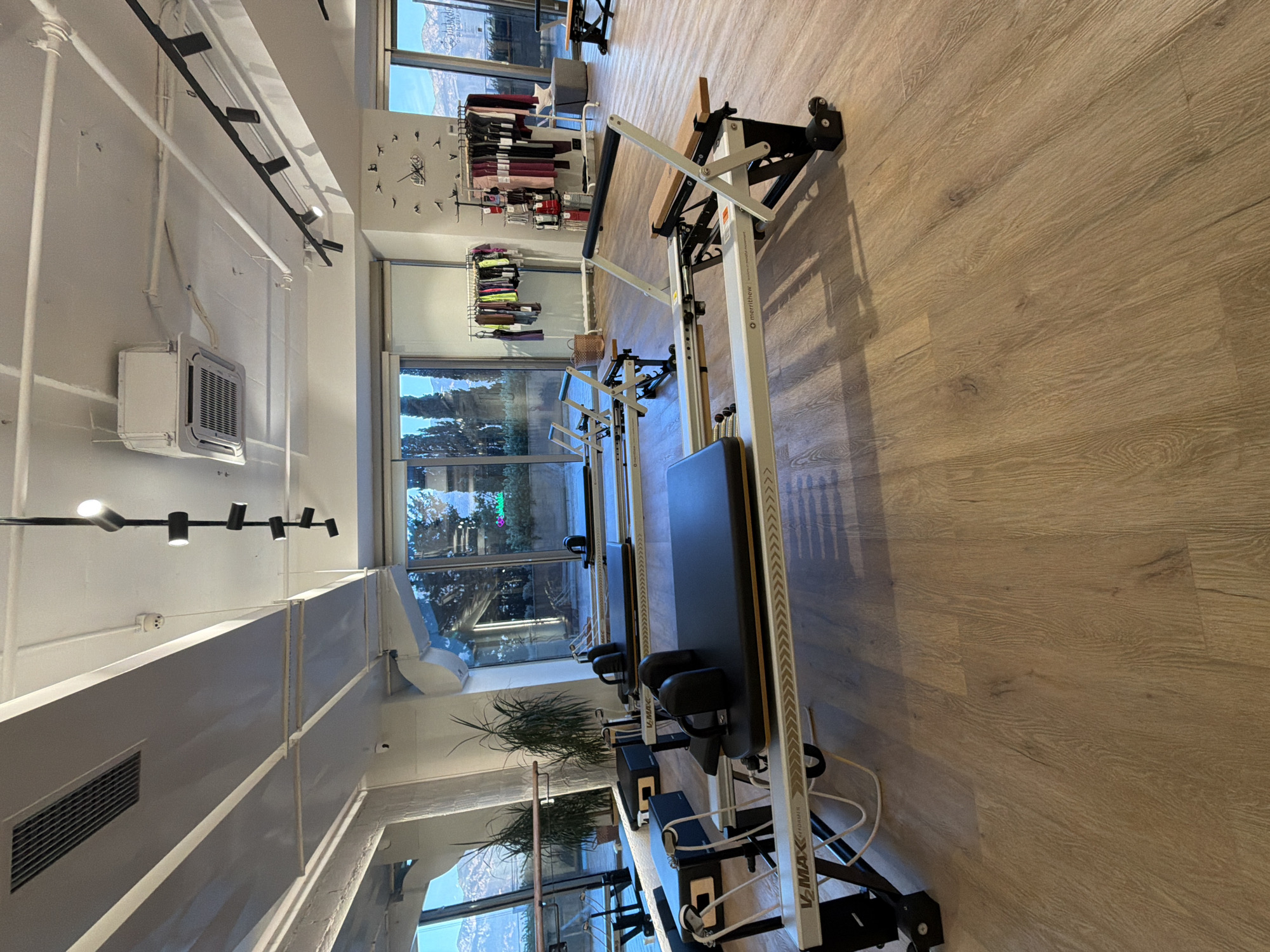 In Boreti, Montenegro, an indoor pilates studio is captured in the image. The room is spacious with a light-colored wooden floor and a white ceiling. Large windows offer a view of trees and mountains. Several pilates reformers, known as V-Max Reformers by Merrithew, are arranged neatly in the studio. These machines are predominantly white and black with some wooden accents. Two reformers are in the foreground, one with its carriage pulled forward, and another further back. Additional reformers are visible in the background. The studio appears well-lit, with overhead lighting and natural light from the windows. A rack of athletic wear, a woven basket, and decorative bird figures on the wall add to the ambiance. The environment is clean and organized, suggesting a professional fitness establishment.
