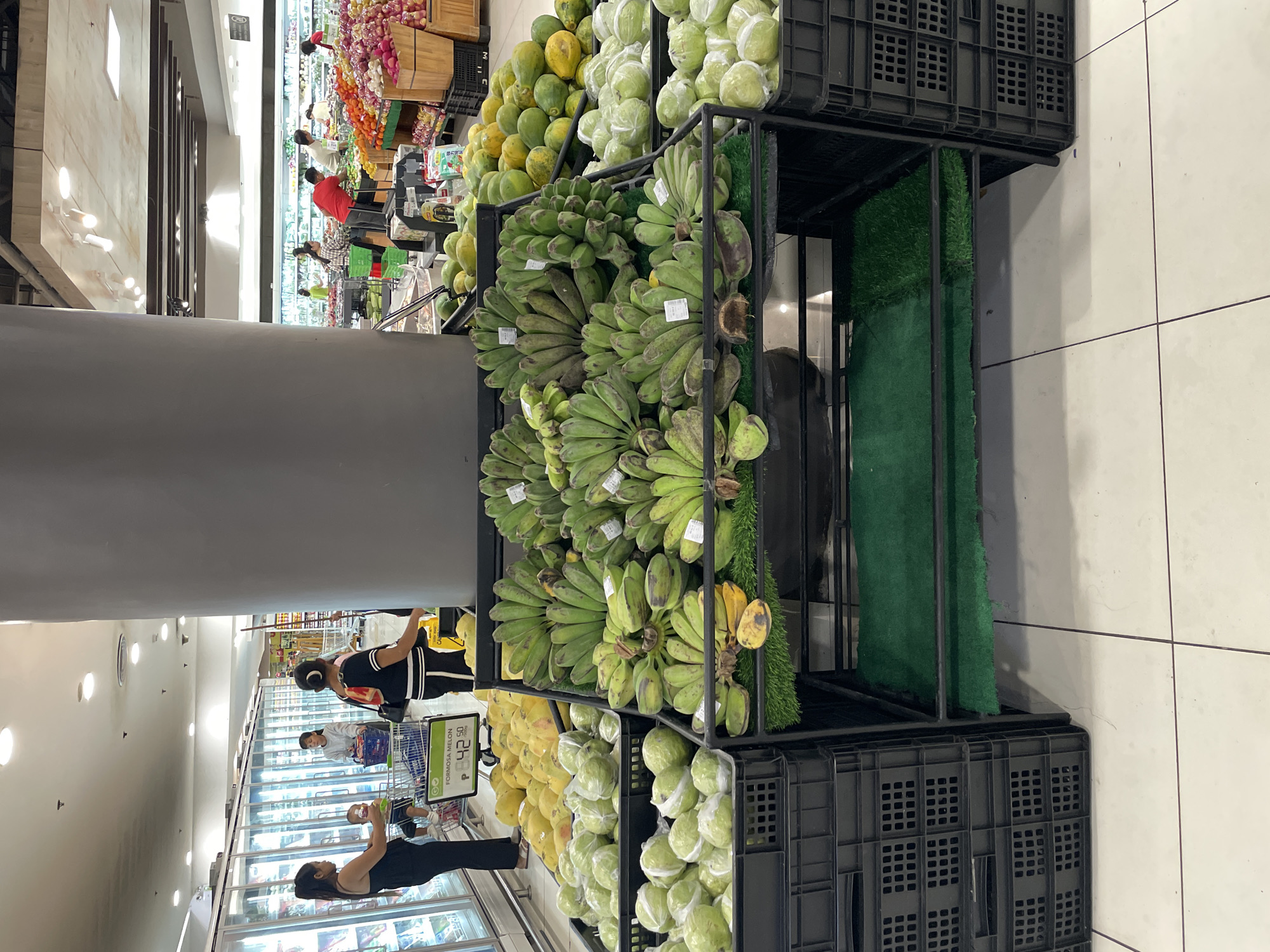 In a supermarket in Davao City, Philippines, a display of fruits is showcased. In the foreground, a large grey pillar stands centrally. To the left of the pillar, in black crates, are piles of guavas, some individually wrapped in clear plastic. To the right of the pillar, on a tiered display with artificial green grass, are bunches of green bananas. Behind the bananas and guavas, a large pile of yellow and green papayas are visible. Further in the background, other shoppers are seen perusing the aisles and interacting with merchandise. The supermarket appears well-lit and stocked with a variety of produce.
