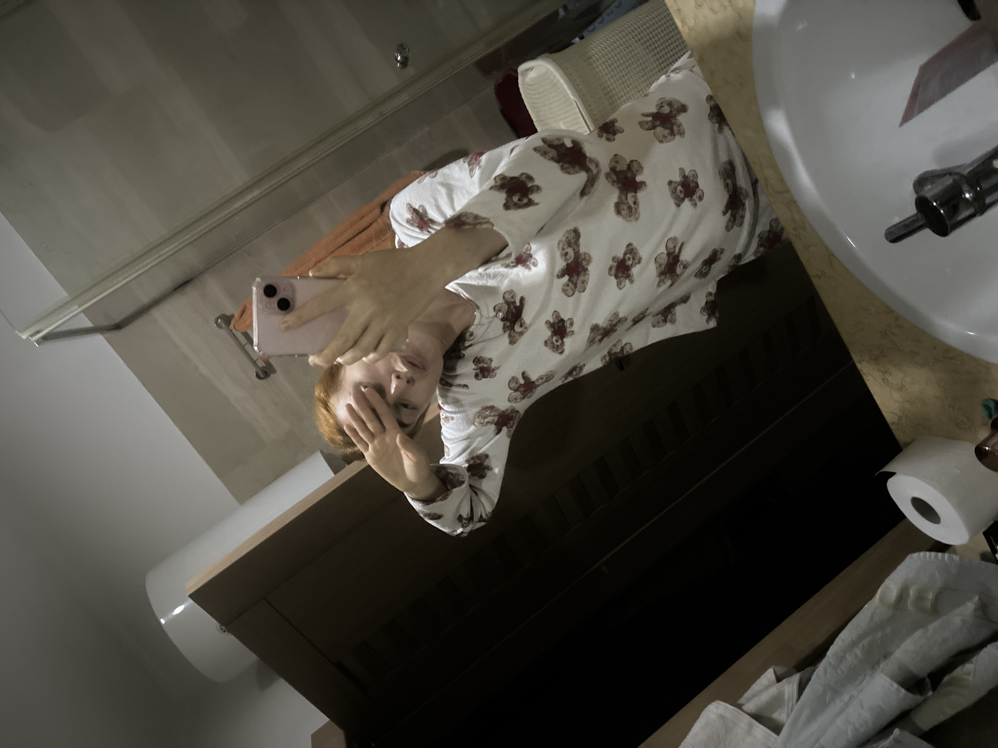 A woman in teddy bear pajamas is taking a selfie in a bathroom mirror. She is holding a pink iPhone. The bathroom has a white sink, a beige countertop, a toilet, and a roll of toilet paper. The location is unknown and cannot be determined to be the city of Budva, Montenegro.