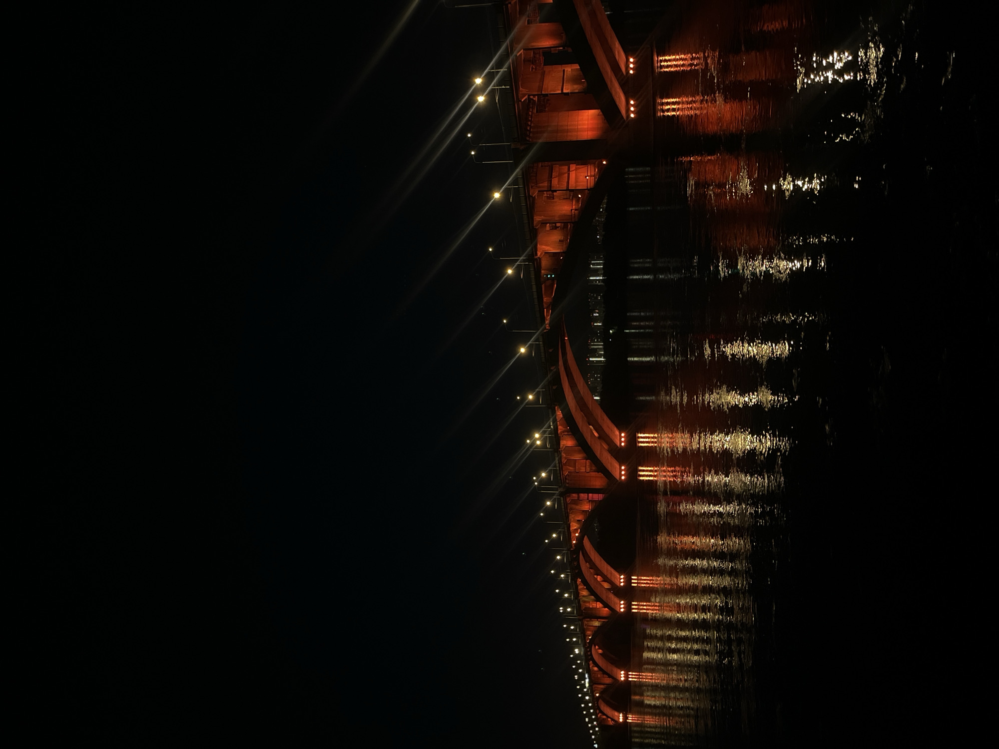The image displays a long, structural bridge illuminated at night, stretching horizontally across a body of water. The bridge structure itself appears to be a reddish-brown or orange hue, accentuated by decorative lighting. Along the top edge of the bridge deck, a series of bright, yellow-white streetlights cast strong beams of light, creating distinct starburst effects against the dark night sky. Additional smaller, orange lights are visible illuminating the underside of the bridge's architectural elements.
The still surface of the water below vividly reflects the illuminated bridge and its lights, forming shimmering, elongated streaks of light that mirror the structure above. Subtle ripples on the water's surface cause these reflections to appear slightly distorted. The sky is pitch black, indicating a clear night with no visible clouds. No people, vehicles, or other activities are discernable on the bridge or in the surrounding environment. The scene is devoid of any visible text. The overall atmosphere is one of serene nighttime beauty, showcasing the illuminated infrastructure. Given the context, this bridge is located in Krasnoyarsk, Russia.