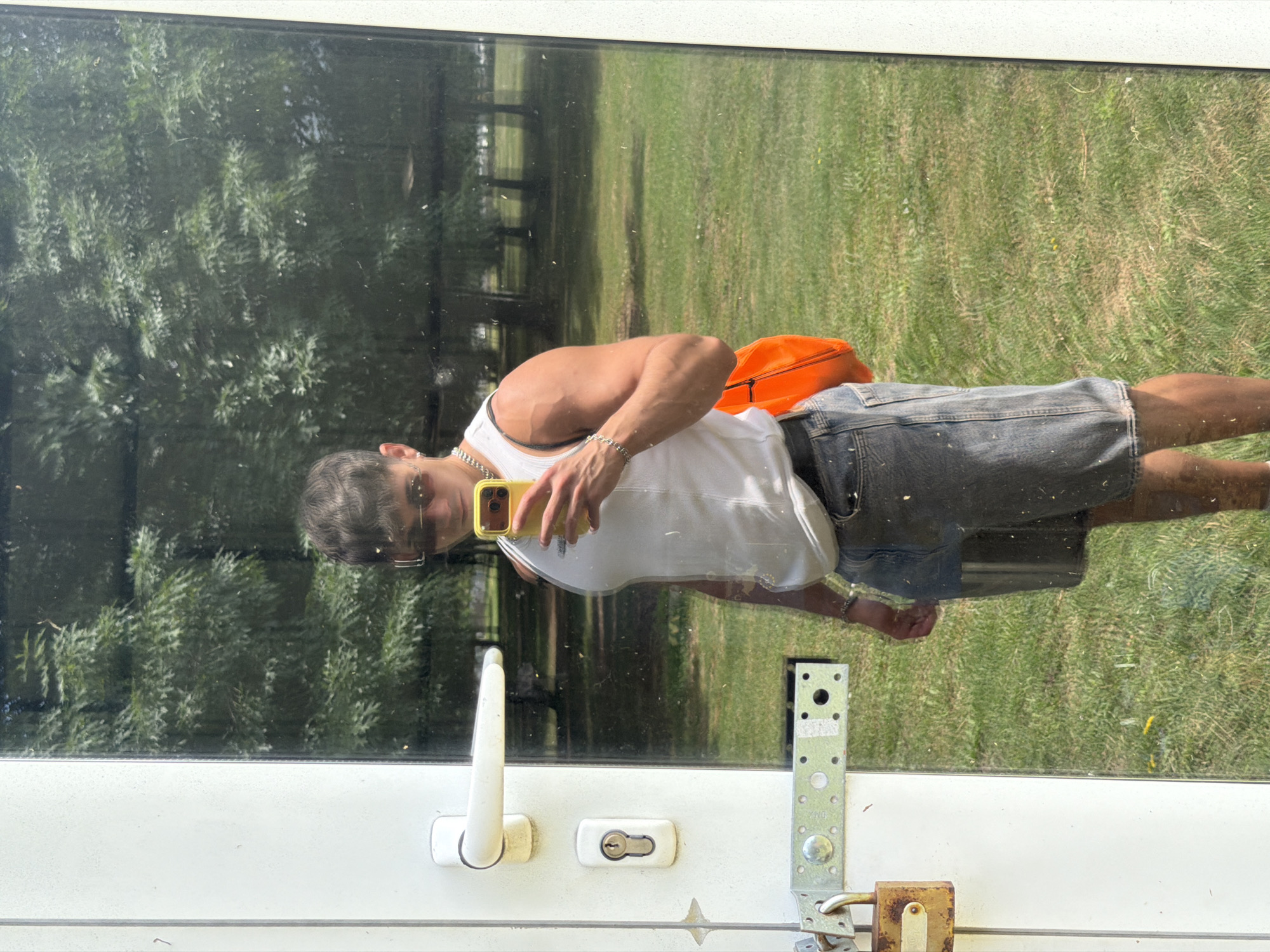 A young man is taking a selfie in a reflection of a glass door, outdoors. He is wearing a white tank top, denim shorts, and sunglasses. He is holding a yellow smartphone in his right hand. The background appears to be a park with trees and grass. The reflection shows a door with a handle, a lock, and a metal bracket with a padlock. The scene is likely in Sofia, Bulgaria, as suggested by the presence of a Sofia brand lock.