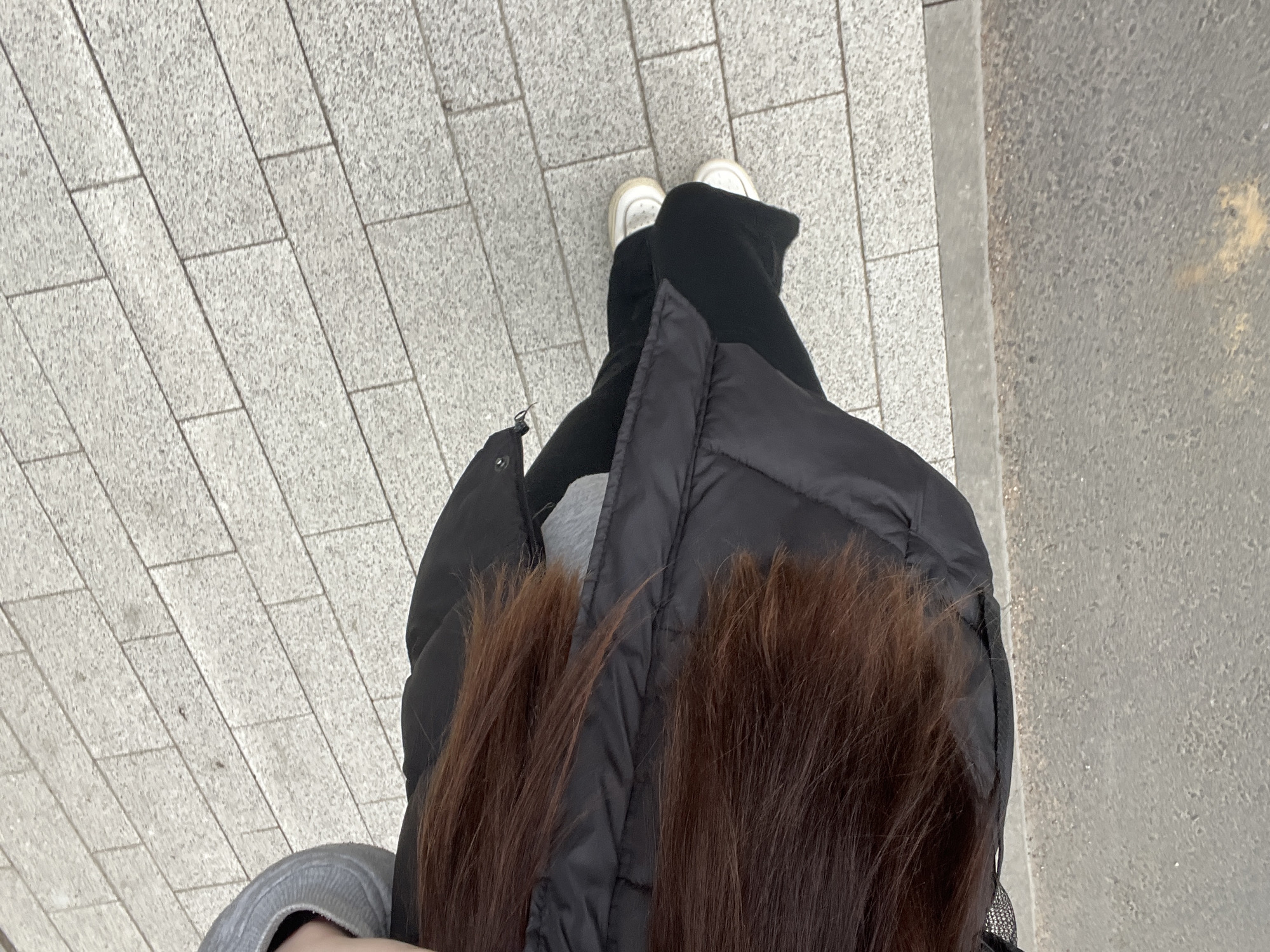 A top-down view captures a person standing on a paved walkway. The walkway consists of rectangular, light grey blocks. The person is wearing white sneakers, black pants, a grey top, and a black puffer jacket. Their long, brown hair falls over the jacket. The scene appears to be outdoors. There is no visible reference to suggest the city of Saint Petersburg, Russia.