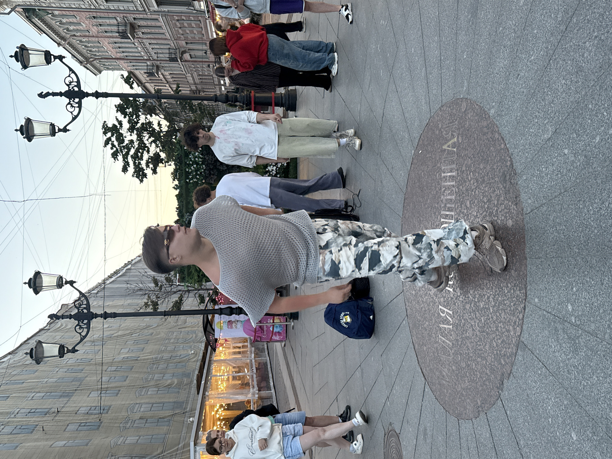 This media file captures an outdoor scene on a paved street lined with buildings. The image is oriented vertically, giving the impression of a sideways perspective. Several people are visible, most of whom appear to be casually strolling or standing around.
In the foreground, a young person in a gray, textured shirt and camouflage pants is striking a pose, with one leg bent and the other extended. They are wearing sunglasses and sneakers. Behind them, a group of people are standing, some in casual attire. One individual wears a red hooded sweatshirt and blue jeans, while another is in a white t-shirt and light-colored pants. Further back, more people are gathered, suggesting a public space or gathering.
Street lamps with ornate black metalwork and glass fixtures are prominent elements in the scene, suggesting it might be evening or dusk, or that they are purely decorative. The buildings lining the street appear to be old, with detailed architectural features and rows of windows, some covered by a protective sheeting. In the background, trees are visible, and the sky appears to be a pale, possibly overcast, hue.
A circular stone inlay on the pavement features engraved text. The visible portion reads "АНЯ". Given the location context of Saint Petersburg, Russia, this could be a street name, monument inscription, or part of an artistic installation. The overall atmosphere seems relaxed and everyday, with no indication of a specific event beyond people enjoying a public space.