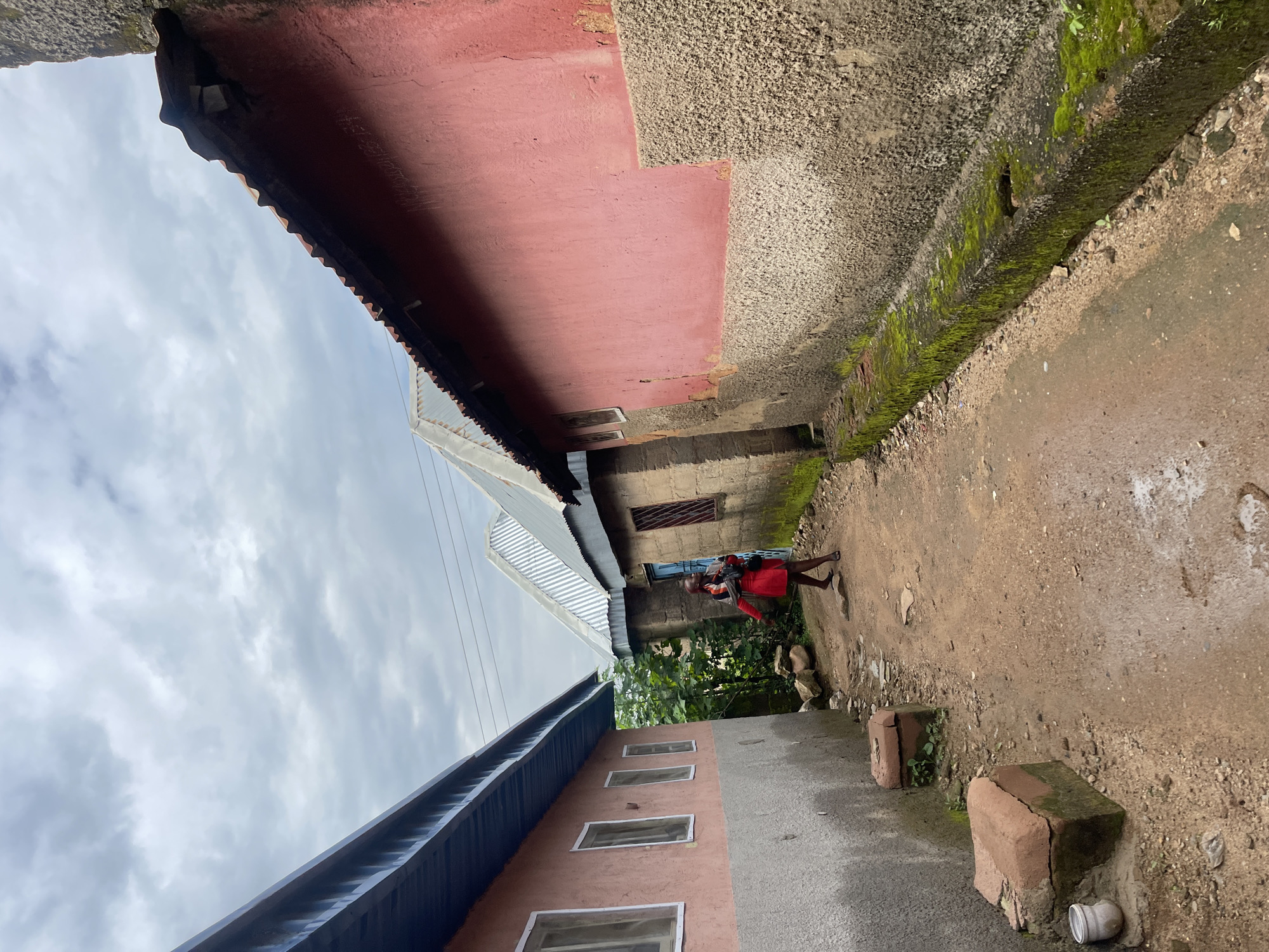 The image shows a narrow dirt path between two buildings. The building on the left is painted pink and gray with several windows. The building on the right is painted pink and gray, with a corrugated metal roof. A person wearing a red dress and carrying a bag is walking away from the viewer down the path. The sky is cloudy. The scene appears to be in an urban environment, likely a residential area in Rafin Par, Nigeria.