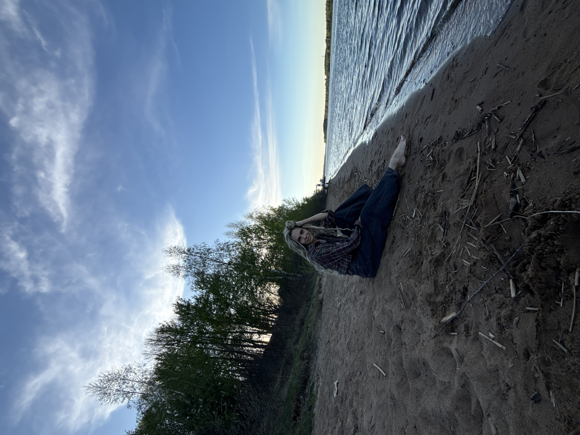 A person lies on a sandy beach with bare feet near a body of water. The individual, likely female, has long blonde hair, wears a plaid shirt and blue jeans, and is positioned at an angle. The background includes a blue sky with white clouds, a line of trees, and water. The foreground shows sand with scattered pieces of driftwood. Location is presumed to be near a body of water in or near Rusanovo, Russia.