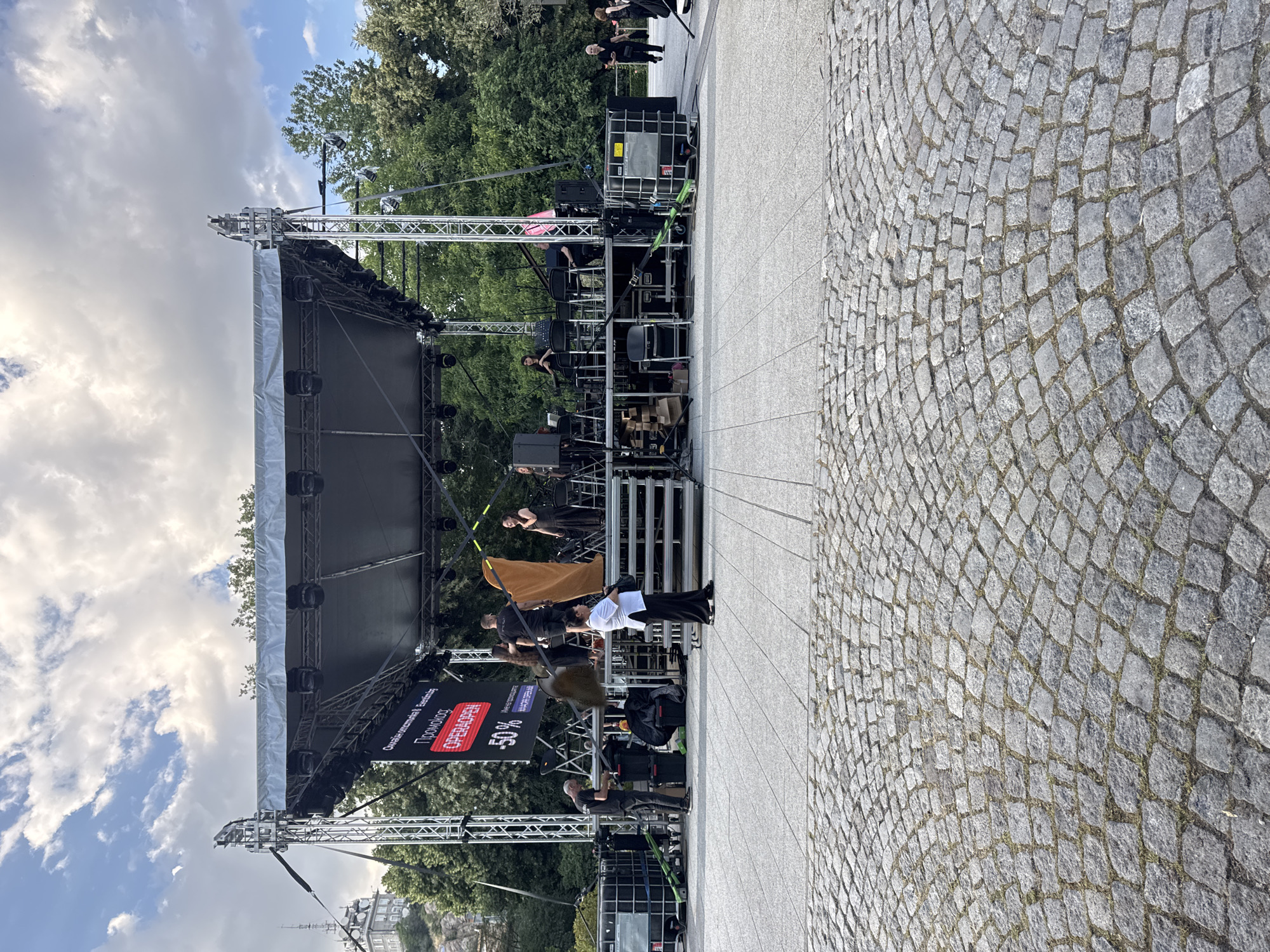 This is an outdoor, eye-level, full shot of a stage setup in Plovdiv, Bulgaria, likely in front of the Ivan Vazov National Library or in the Tsar Simeon's Garden, given the architectural style of the building visible in the background and the overall urban park setting.
The scene is dominated by a large, temporary stage structure with a black roof supported by silver metal scaffolding. Several spotlights are affixed to the underside of the roof. A large digital screen on the stage displays text in Cyrillic and Latin alphabets, advertising "OPERAOPEN" with "-50%" and other promotional text.
On and around the stage, several individuals are present. Some appear to be setting up or managing equipment. One person in a white shirt and black pants is standing in the foreground on the paved area, facing away from the camera. Others are visible on the stage, including a woman in a black dress and others in dark clothing. There are musical instruments and chairs set up on the stage.
To the left of the stage, large black speaker cabinets or equipment cases are visible. The ground in the foreground is paved with cobblestones arranged in a curved pattern. Behind the stage, trees form a dense green backdrop, and a building with a distinct spire or tower is visible on a hill in the distance. The sky above is partly cloudy with a mix of blue sky and white clouds. The overall atmosphere suggests preparation for an outdoor performance or event.
