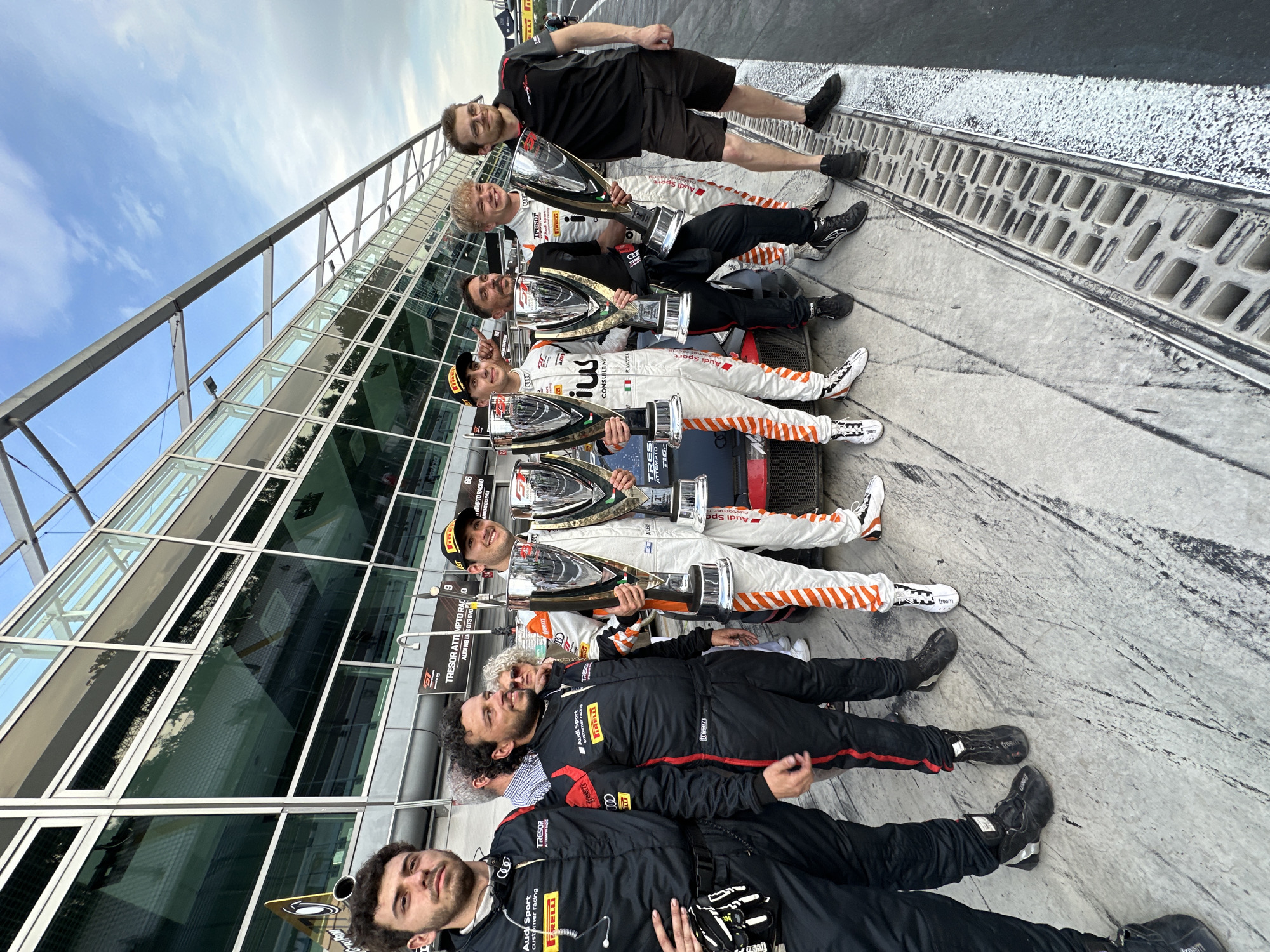 A group of nine people are lying on the ground, holding trophies, in what appears to be a celebratory photo following a race. The location is in Monza, Italy, based on the signage and overall environment.
The individuals are aligned from bottom to top in the frame. The bottom two individuals are wearing black racing suits with red accents and appear to be part of the support team. The next five are wearing white racing suits with Italian flag insignias, logos, and some orange detailing. They each hold large silver trophies with black bases. The top two individuals are wearing similar racing attire, but one is standing, and the other is lying down in what appears to be a support position.
A building with a glass facade and a steel frame is in the background. The ground is a gray concrete surface, and a drain runs along the edge. The sky is visible in the background, suggesting an outdoor setting.