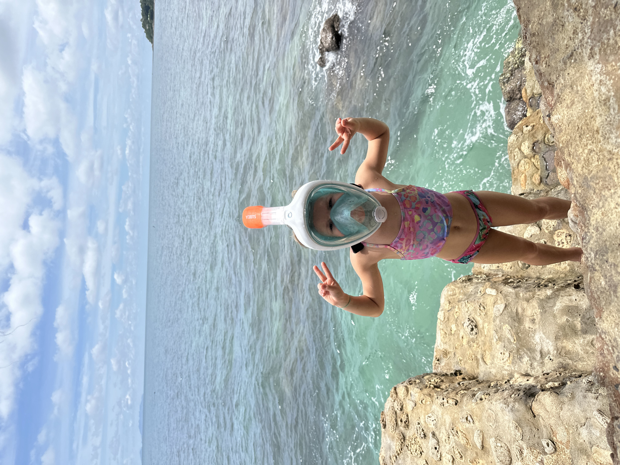 In ตำบลเกาะพะงัน, Thailand, a young girl stands on a rocky outcrop overlooking the ocean. She is wearing a full-face snorkel mask and a colorful swimsuit. Her hands are raised, with her index and middle fingers extended in a peace sign on each hand. The water is a clear turquoise color, and the sky is filled with scattered white clouds. The rocky area the girl is standing on appears to be a natural formation with some rough, weathered stone steps. The background shows the expanse of the ocean and a distant, tree-covered island.