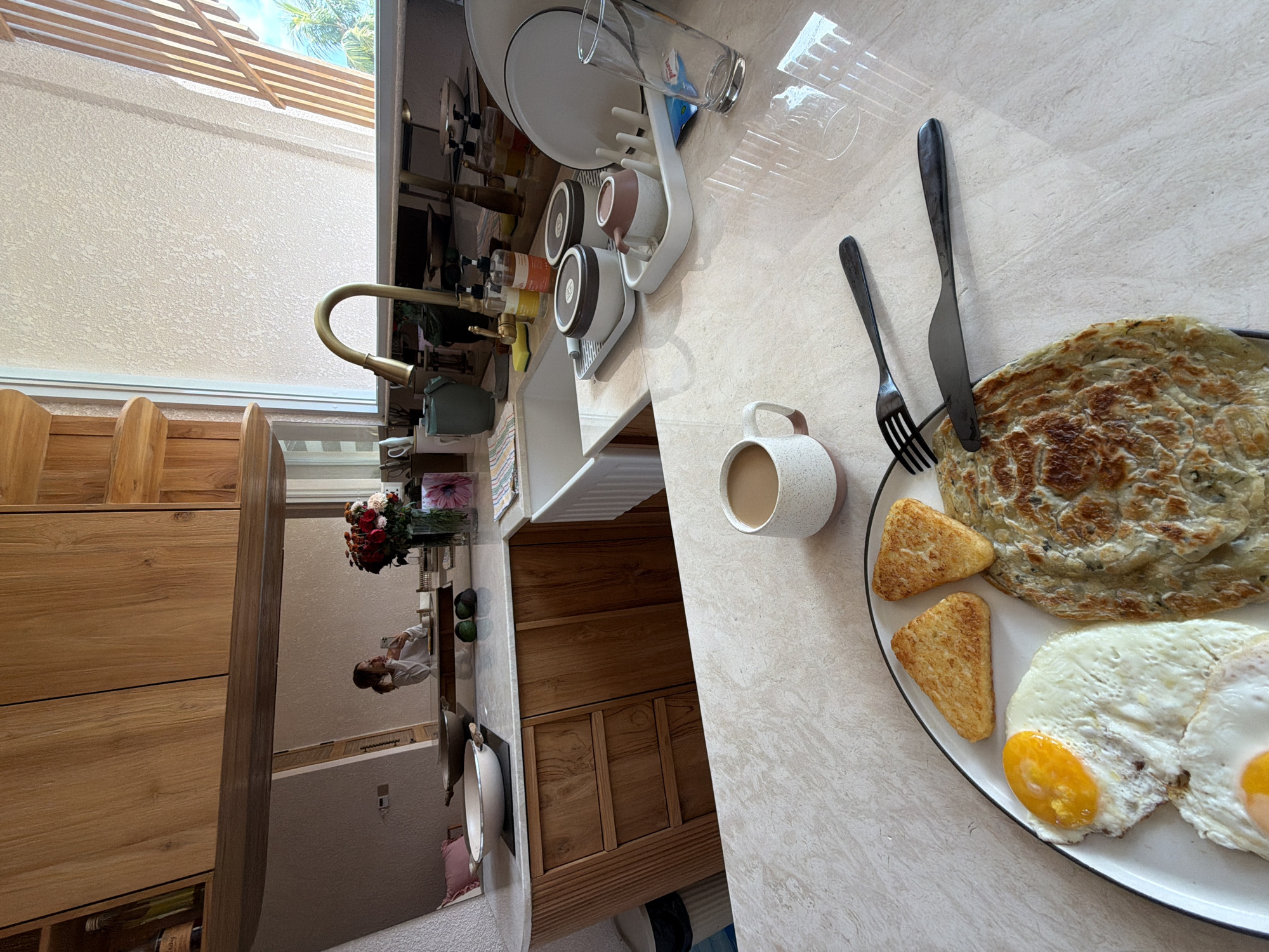 A rotated eye-level shot reveals a breakfast plate on a marble countertop, likely in a kitchen. The breakfast consists of two sunny-side-up eggs, two hash browns, and a roti-like flatbread. A fork and knife rest on the plate. To the left, a speckled mug is half-filled with a light-colored liquid, potentially coffee or tea. The countertop reflects overhead sunlight.
Behind the breakfast scene, a kitchen sink with a faucet and a dish-drying rack is visible. The rack holds plates, mugs, and a glass. Further back, a wooden cabinet with drawers and a shelf containing wine bottles is present. A mirror reflects a person wearing a white shirt, along with a vase of flowers. Outside the window, a pergola structure is apparent, with a palm tree visible in the distance.
The location is plausibly in Mengwi, Indonesia, given the tropical vegetation visible through the window, which suggests a tropical or subtropical climate typical of that region. However, this is based solely on visual cues and lacks definitive proof.