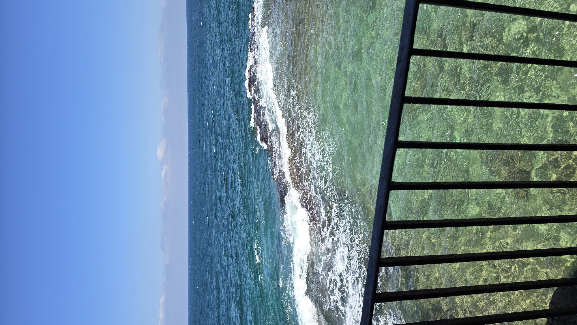 A vertical, eye-level shot captures a view of the Mediterranean Sea from behind a black metal railing. The railing, consisting of vertical bars and a horizontal top bar, occupies the foreground. Beyond the railing, the water transitions from deep teal in the distance to a clear, shallow greenish hue closer to the shore. Waves crash against a rocky outcrop jutting into the water. The sky above is a clear, bright blue with a few wispy white clouds forming a thin band near the horizon. The scene is likely located along the coast of the city of Acre, Israel, known for its historical port and clear waters.