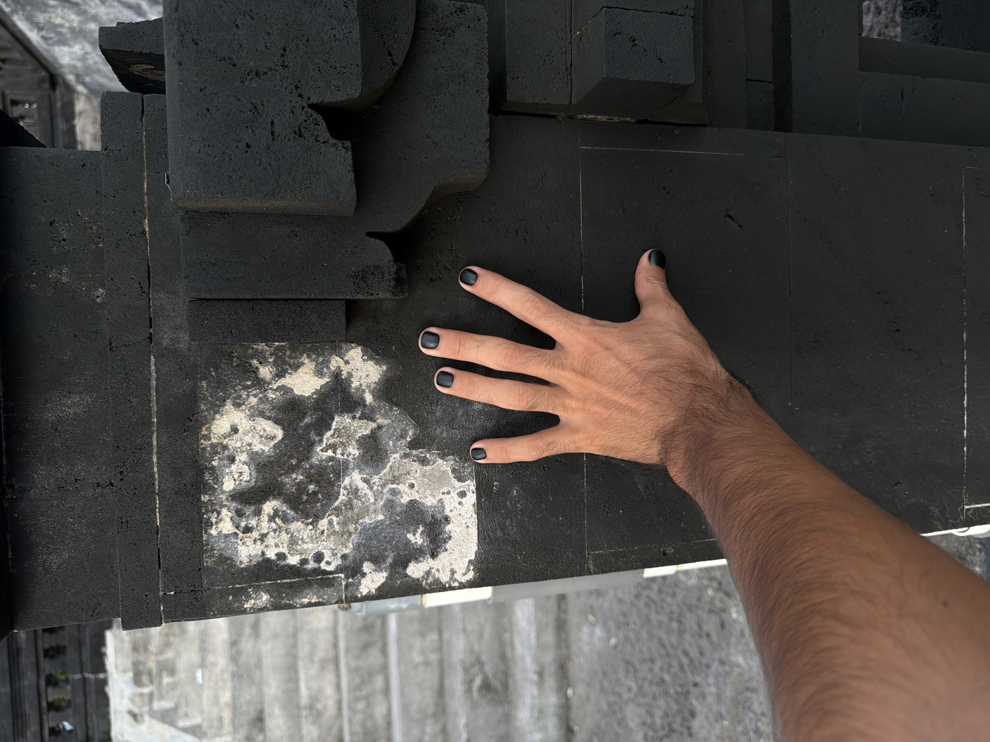 This high-angle, close-up shot, taken in North Kuta, Indonesia, features a person's right arm and hand resting on a dark, textured surface. The hand is positioned palm-down, with all five fingers spread. Each fingernail is painted with matte black nail polish, creating a striking contrast against the medium-toned skin. The arm, visible up to the forearm, is covered in fine, dark hair.
The surface beneath the hand is composed of various dark grey to black, block-like structures, possibly architectural elements or an abstract sculpture made of concrete or a similar rough material. These blocks exhibit a coarse, porous texture and some irregular, chiseled edges. To the lower left of the hand, a portion of the surface is distinctly different, featuring a mottled pattern of white, light grey, and dark grey, suggesting weathered concrete or stone with lichen or paint residue. This lighter section creates a strong visual division with the darker blocks.
In the background, below the main surface, out-of-focus grey concrete steps or a similar structured ground are visible, reinforcing an outdoor setting. The bright, even lighting indicates daytime conditions. There is no visible text or explicit emotional expression, but the image carries a minimalist and textural aesthetic, highlighting the interaction between the human element and the stark environment.