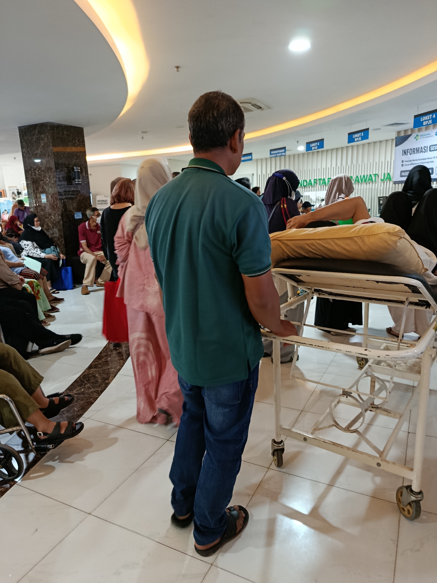 The image depicts an interior scene, likely a waiting area in a hospital or clinic in East Jakarta, Indonesia. A man in a green t-shirt and blue jeans stands with his back to the camera, facing a stretcher on wheels. On the stretcher, a person lies down, covered by a cream-colored sheet. The environment includes a tiled floor with a decorative patterned inlay, a dark, polished pillar, and a curved ceiling with yellow lighting. In the background, a line of people are waiting, some seated and others standing. Signs indicating "LOKET" (ticket window) and "INFORMASI" (information) are visible on the wall, suggesting a formal registration or service area. One person in the background is pushing a wheelchair. The overall atmosphere suggests a busy, medical-related environment.