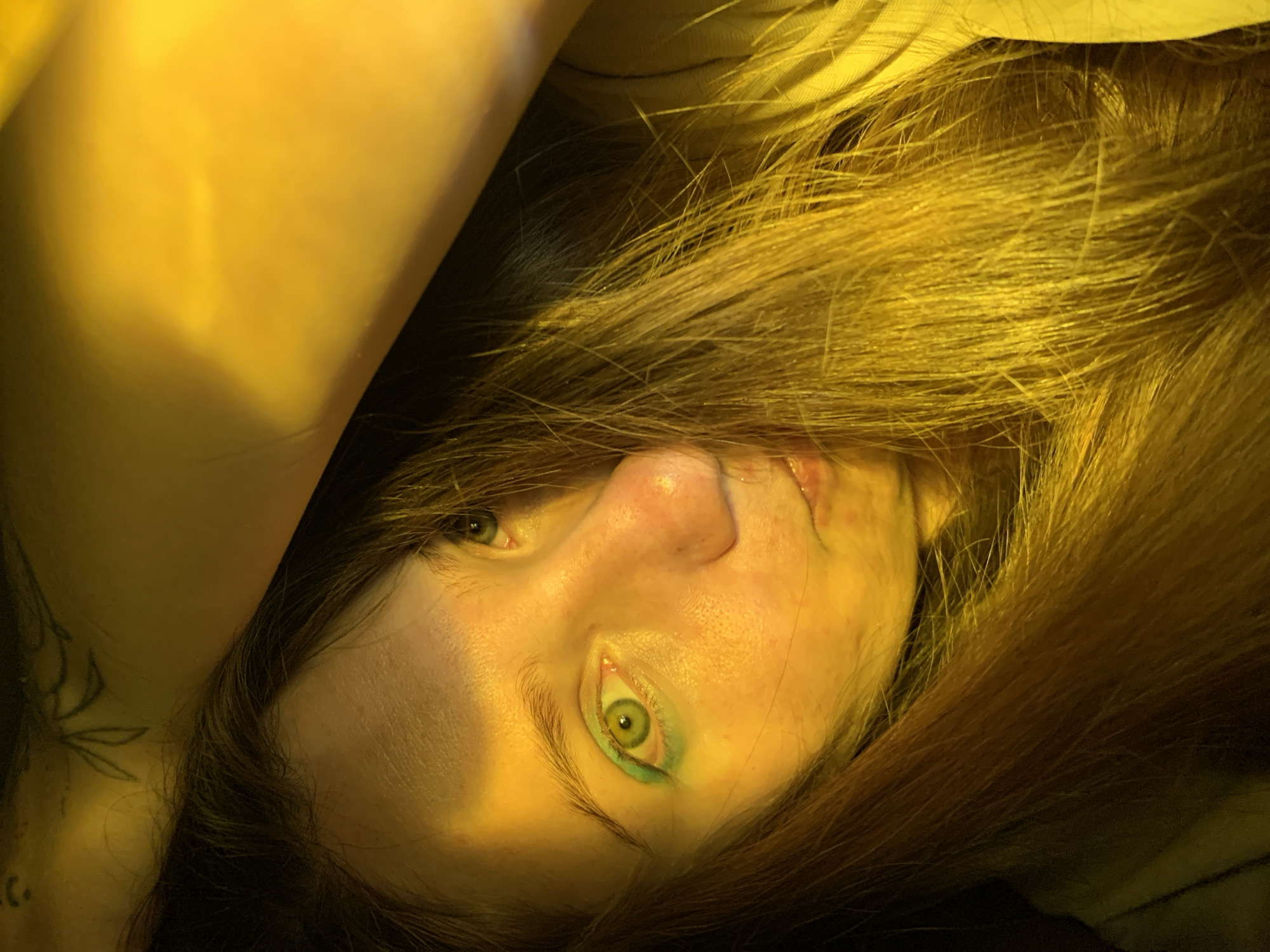 A young person's face is framed by long, dark hair. The subject has green eyes and is wearing green eyeshadow. The lighting in the image is predominantly yellow, casting a warm glow on their skin. A tattoo of a plant with leaves is visible on the upper part of the image. The scene appears to be indoors, with a hint of a striped pattern on the fabric in the upper right corner. The overall impression is intimate and artistic. There is no information available to determine the specific location in the city of Chaikovsky, Russia.