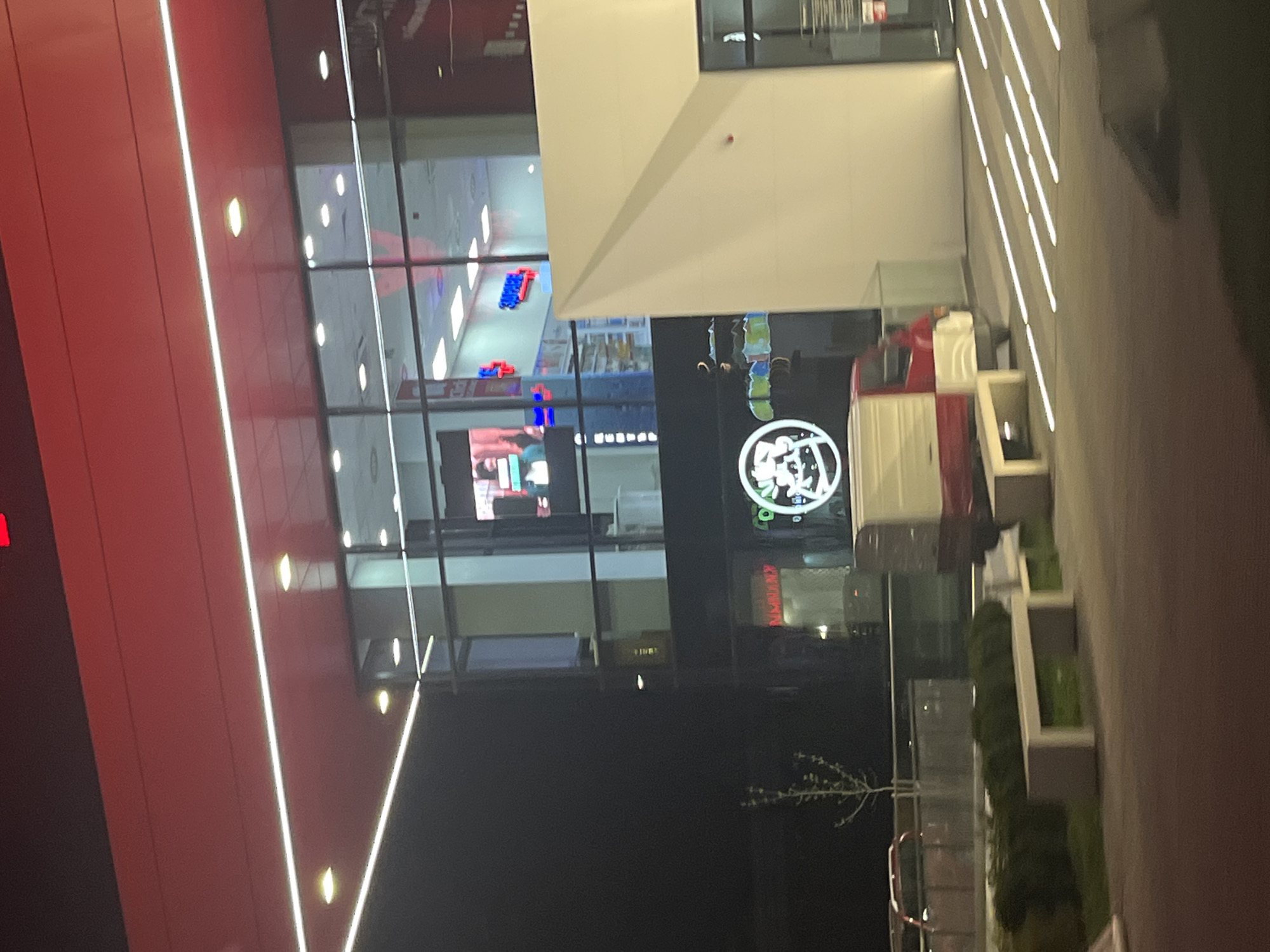 The image shows a nighttime view of a modern building exterior. The building features a red facade with vertical white light strips and large glass windows reflecting interior lights and signs. A bright white structure juts out from the building. Reflected in the glass are signs, including a circular logo with text and images. A red-and-white structure, possibly a decorative element, sits on the sidewalk in front of the building, adjacent to landscaping with green plants in concrete planters. A bare tree is visible near the sidewalk. The location appears to be in Sofia, Bulgaria.