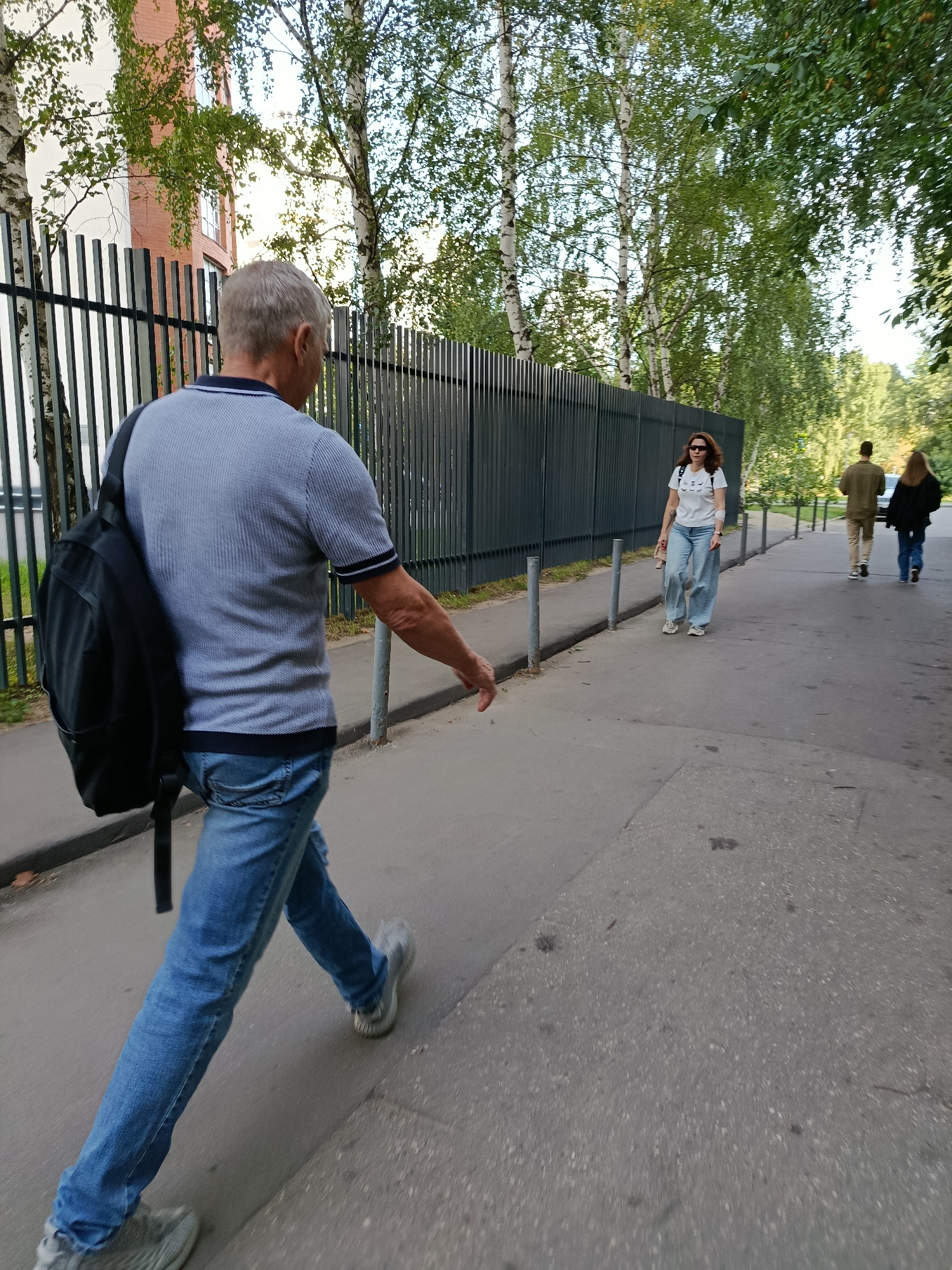 In Moscow, Russia, on a pedestrian sidewalk next to a dark metal fence, a middle-aged Caucasian man with graying hair and a blue polo shirt walks away from the camera with a black backpack on his left shoulder. He is wearing blue jeans and gray sneakers. Further down the sidewalk, a Caucasian woman with dark hair, sunglasses, and a white t-shirt with text and loose-fitting jeans, walks towards the camera. Behind her, a man in a brown jacket and tan pants and a woman in a black jacket are walking away from the camera. A multi-story building with light-colored brick and windows is visible behind the fence, partially obscured by trees. The scene is set on a sunny day with deciduous trees lining the sidewalk.