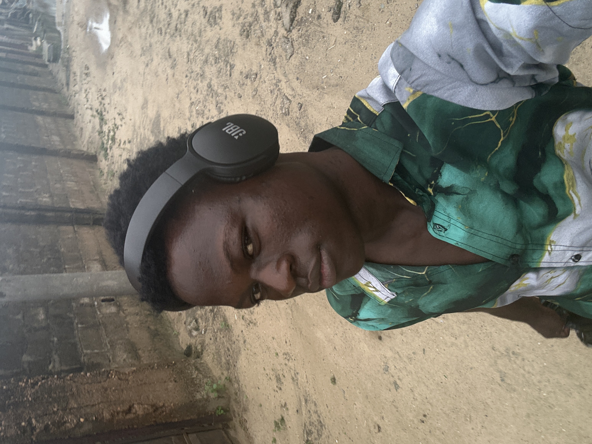 A young person with dark skin and short, dark hair is wearing black JBL headphones and a green and gray shirt with a marbled pattern. The person is looking downwards and slightly to their left, with a neutral expression. The background consists of a sandy or dirt ground and a low wall made of rough stone or concrete blocks. The lighting suggests it could be daytime, possibly overcast or in shade, as there are no strong shadows. The image is oriented vertically, with the person's head at the top of the frame. The setting appears to be an outdoor, possibly rural or semi-urban area in Ajara, Nigeria.