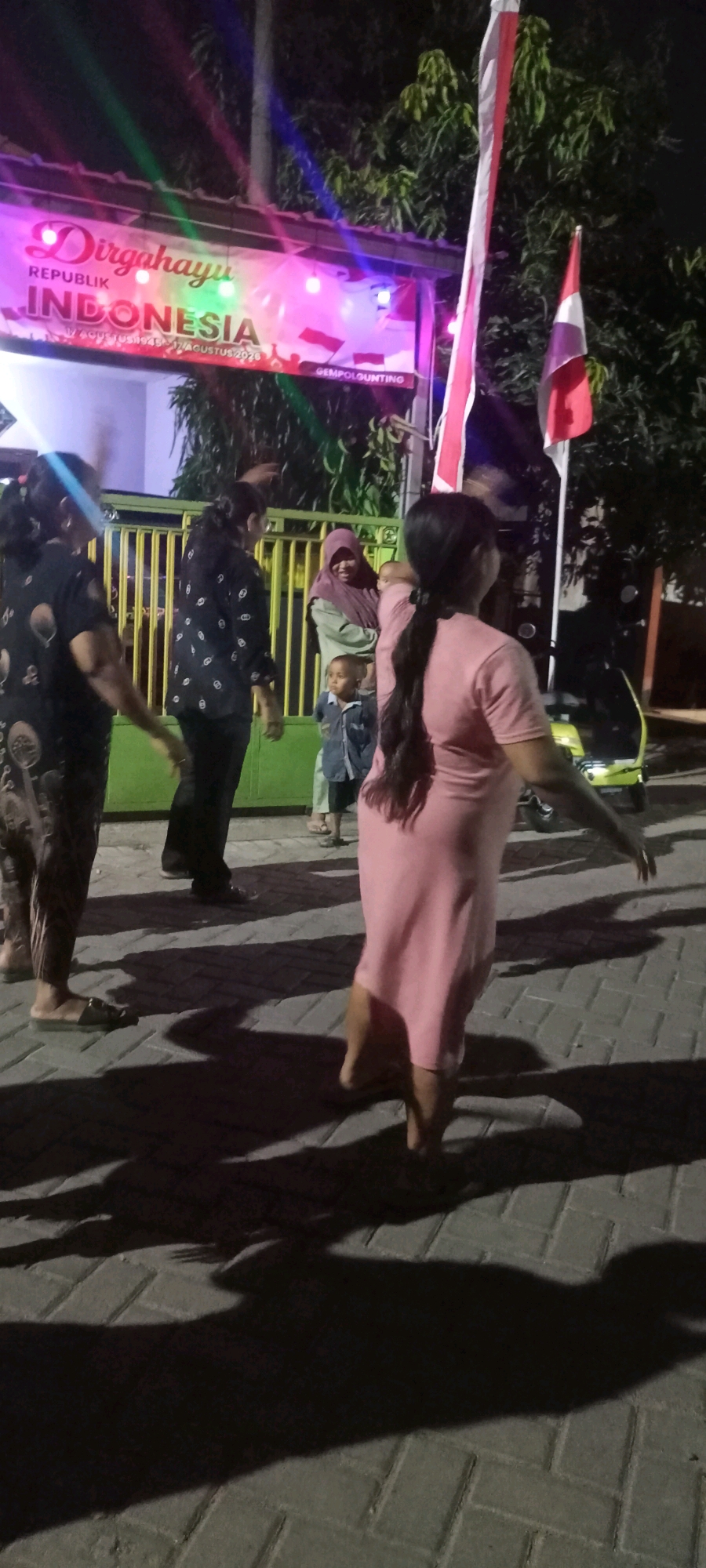 The image shows an outdoor scene at night, likely in the city of Gempolklutuk, Indonesia, based on the banner visible. The banner, illuminated with pink and green lights, reads "Dirgahayu REPUBLIK INDONESIA" and displays dates including "17 AGUSTUS 2020". Several people are present, with a focus on the right side of the frame on a woman in a pink dress, viewed from behind, with her right arm raised. To her left, a man in a black shirt with a white pattern is visible, also with his arms raised. Further left, another woman in a patterned black shirt is seen from the side, with her left arm raised. In the background, behind a yellow-barred fence, a woman wearing a hijab is holding a baby, and a small child in a blue shirt is standing. A red and white Indonesian flag is prominently displayed on a pole. The ground is paved with interlocking bricks, and shadows are cast on the surface, indicating artificial lighting. A yellow scooter is parked in the background on the right. The overall atmosphere suggests a celebration or gathering.