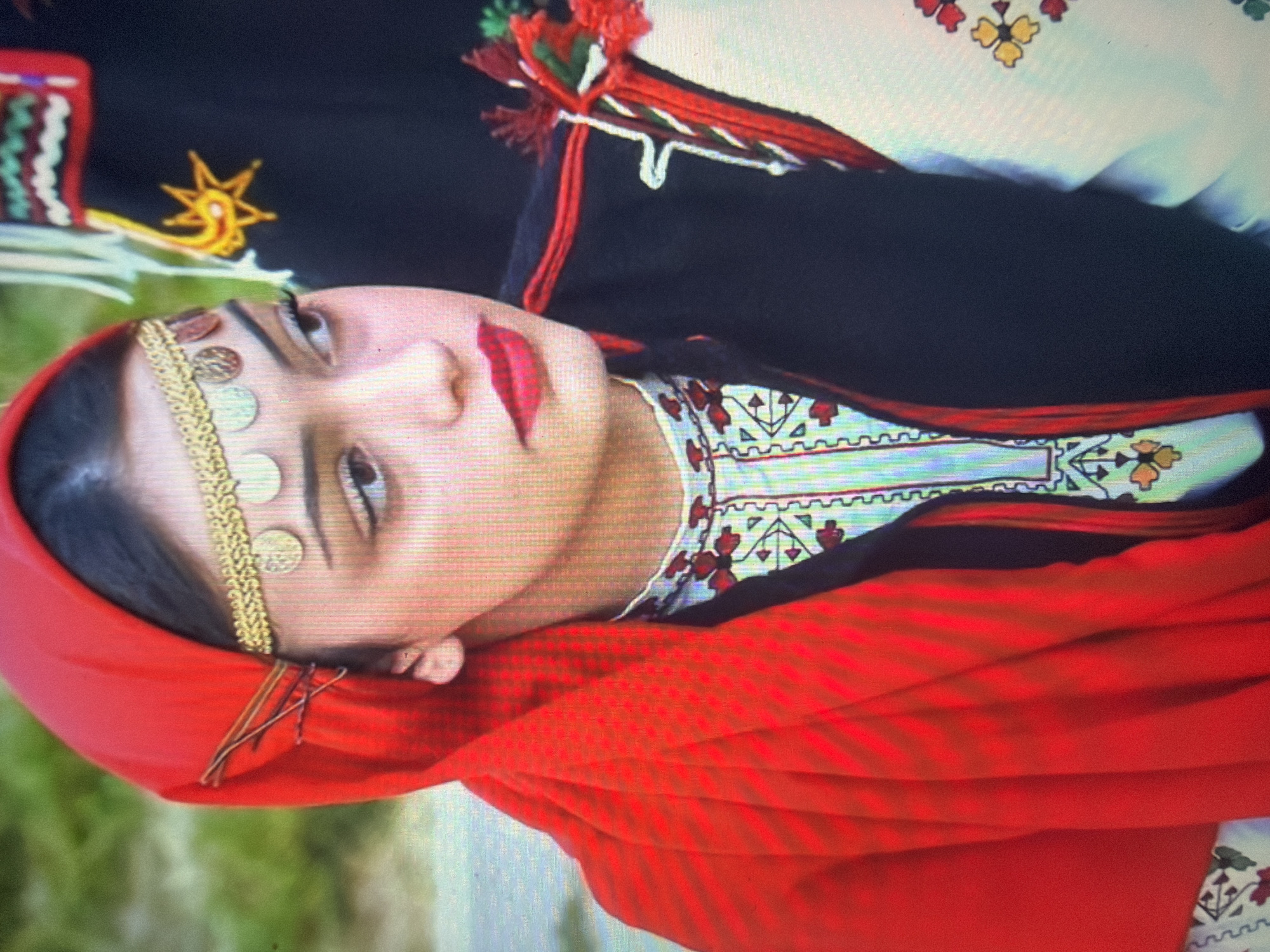 The image depicts a young woman in traditional attire, photographed outdoors in Shumen, Bulgaria. She has dark hair, brown eyes directed towards the right side of the frame, and wears red lipstick. Her head is covered by a bright red headscarf, which is textured and draped over her left shoulder. A gold headpiece, consisting of a braided band adorned with five circular gold coins or medallions, rests across her forehead. She is wearing a white garment with a high collar, intricately embroidered with red, white, and yellow geometric and floral patterns. Over this, she wears a dark (likely black) sleeveless vest with red trim, and decorative red and white braided elements with tassels are visible on her right shoulder. The background includes blurred green foliage on the left. On the right, a dark fabric, possibly part of another individual's traditional clothing, displays a prominent yellow embroidered starburst or sun motif, accompanied by green and red zigzag patterns at its upper edge.