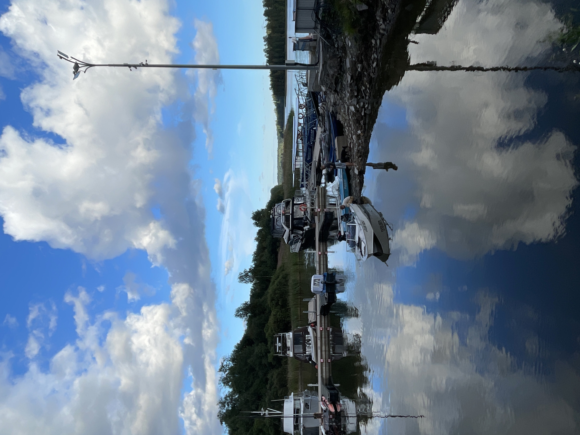 A wide shot captures a serene marina in Berozovo, Russia, under a sky filled with scattered white clouds against a bright blue backdrop. The water in the foreground reflects the sky and the surrounding environment, creating a mirror-like effect. Several boats are docked, ranging from small recreational vessels to larger houseboats and passenger boats. Some of these boats are moored along a wooden dock, while others are positioned closer to the shore. The shoreline is rocky and partially covered with vegetation. A tall lamppost stands on the right side of the frame, its reflection visible in the water. In the distance, a tree-lined bank forms the backdrop to the marina. The scene suggests a calm and peaceful day at the harbor.