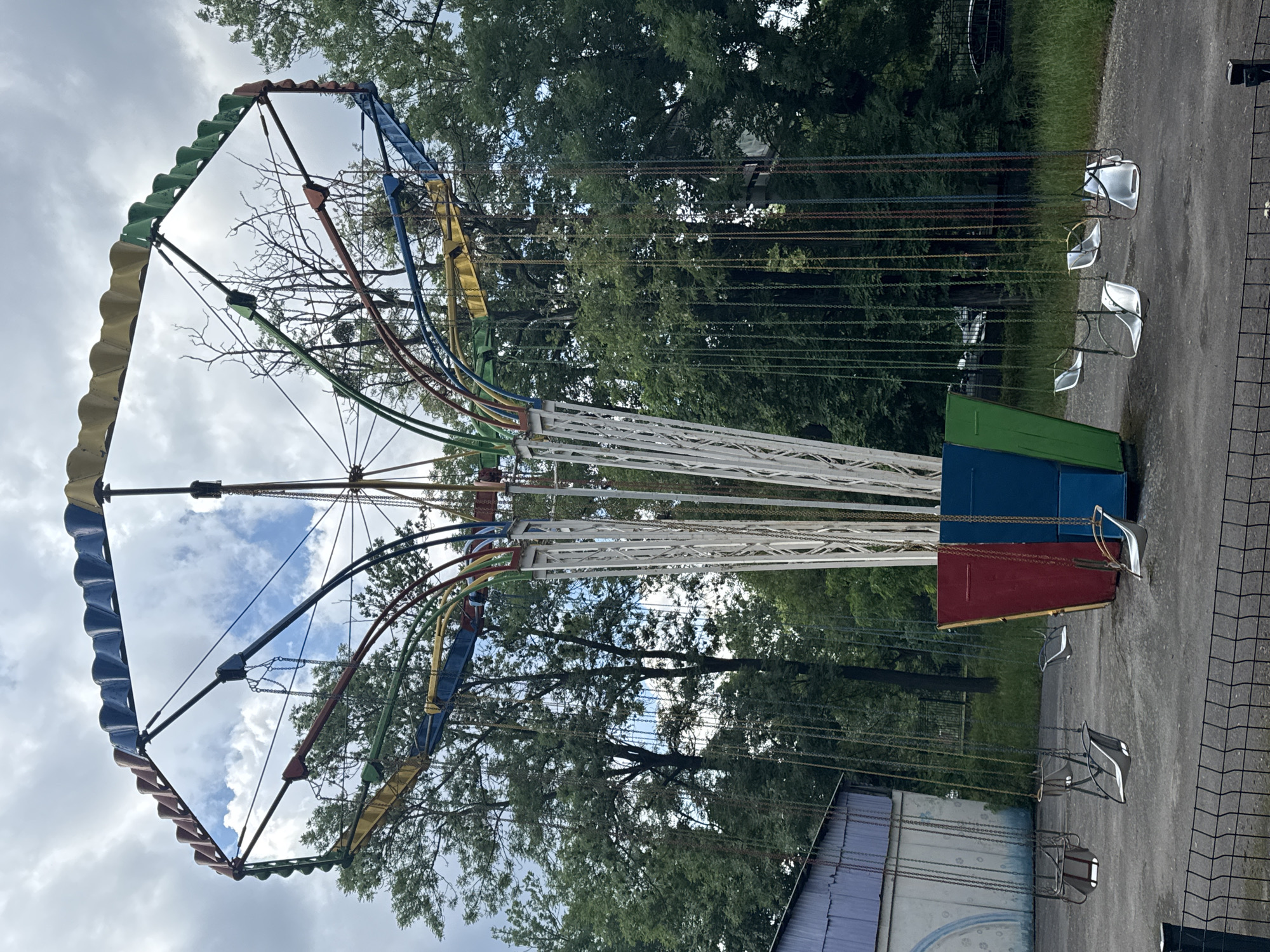 An abandoned amusement park swing ride stands in Vinnytsia, Ukraine. The ride features a central white structure with colorful curved supports that hold up a hexagonal top. Multiple empty swings hang from chains, suspended by the ride's structure. The ground around the ride is paved, with some areas appearing wet. To the left of the ride is a building with a blue roof and white walls, partially covered by peeling paint. The background is filled with lush green trees, with some bare branches visible. The sky is overcast with patches of blue showing through the clouds. A black metal fence runs along the foreground of the image. The scene appears deserted and neglected, suggesting the park has been out of operation for some time.