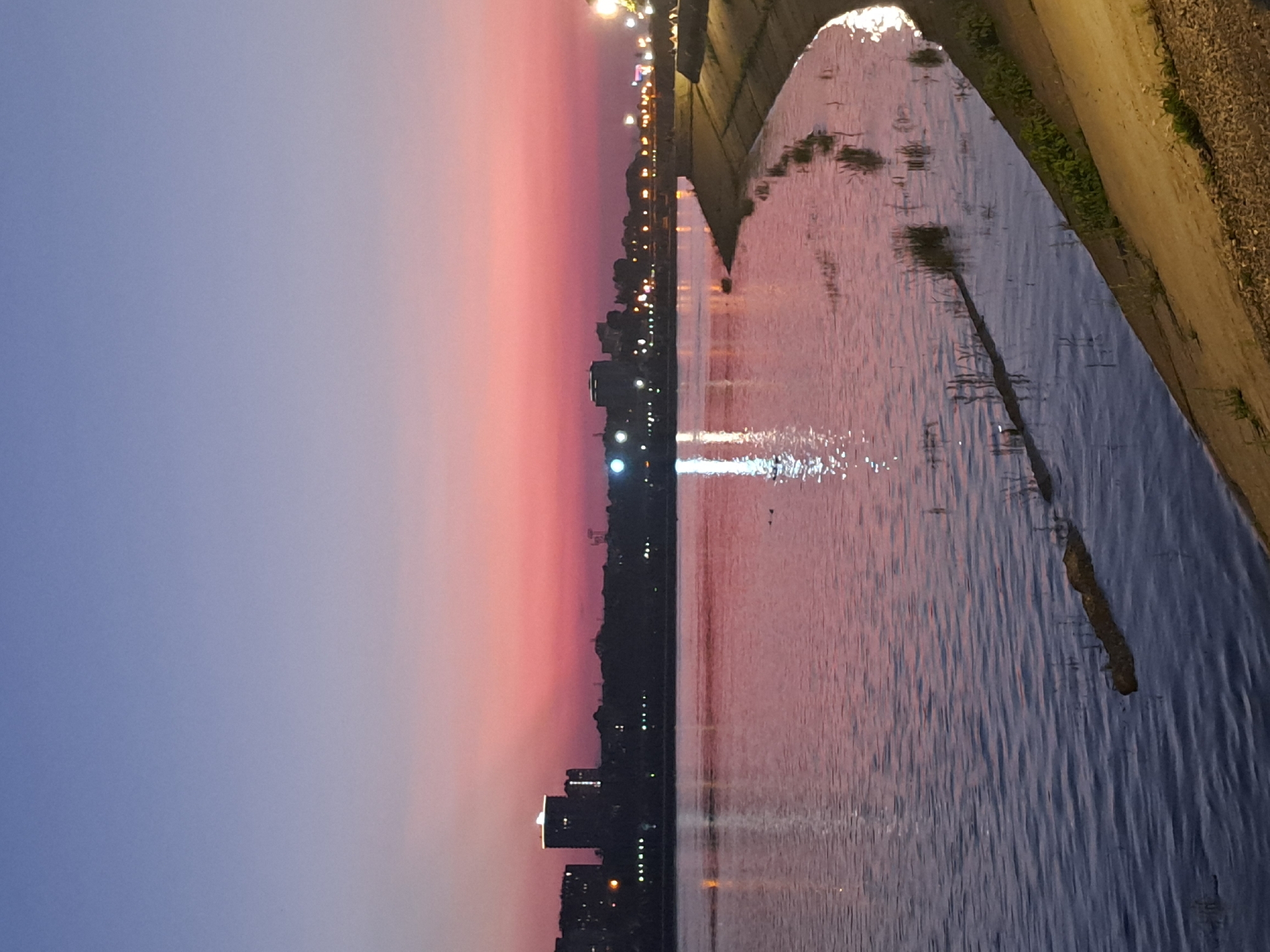 The image depicts a tranquil sunset scene over a body of water in Irkutsk, Russia, likely the Angara River or one of its reservoirs. The sky transitions from a deep blue at the top to vibrant pink and orange hues near the horizon, creating a warm and serene atmosphere. Silhouetted against the colorful sky is the city skyline, with buildings and lights reflecting on the calm water's surface. A concrete embankment lines the right side of the image, illuminated by streetlights, with some vegetation growing along its edge. The water itself is a deep blue, disturbed by gentle ripples and reflecting the colors of the sunset. There are no people visible in the frame, and the overall impression is one of peace and natural beauty.