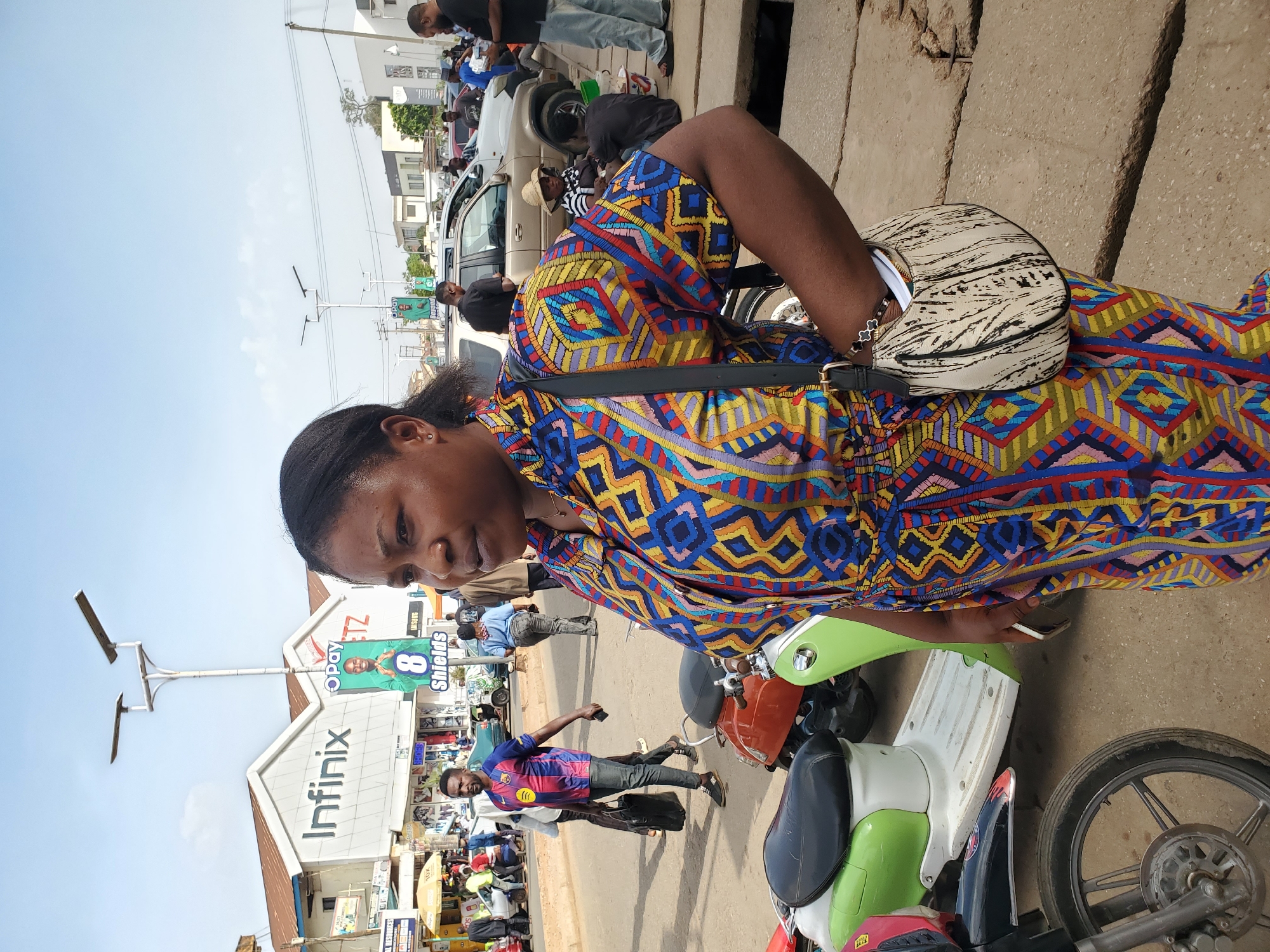 This image captures a bustling street scene in Jos, Nigeria, during what appears to be a bright, sunny day with a clear sky.
The primary subject is a Black woman positioned in the foreground, seen from the waist up. She is wearing a vibrant, multi-colored dress with intricate geometric patterns in shades of blue, yellow, red, orange, and black. A light-colored, crescent-shaped shoulder bag with black abstract markings is slung across her body. Her hair is pulled back, and she is looking downwards and slightly to her right, appearing focused on something in her hand, possibly a mobile phone, or an object near the ground. She stands beside a green and white scooter or small motorcycle.
The background reveals a busy urban environment. Numerous people, predominantly men, are visible walking, standing, and interacting along the street. One man in a blue and red striped football jersey and dark trousers is walking in the mid-ground. Cars and other motorcycles are parked and scattered throughout the scene. Buildings line the street, suggesting a commercial or residential area.
Notable details include a prominent building with a large "Infinix" sign. Adjacent to it, a banner displays "OPay" along with the text "8 Shields" and an image of a person. Part of a "SAMSUNG" sign is also visible further down. Modern streetlights with solar panels are installed along the roadside. The ground consists of a mix of paved and unpaved surfaces, characteristic of a busy street or marketplace. The overall impression is one of everyday urban life and commerce.