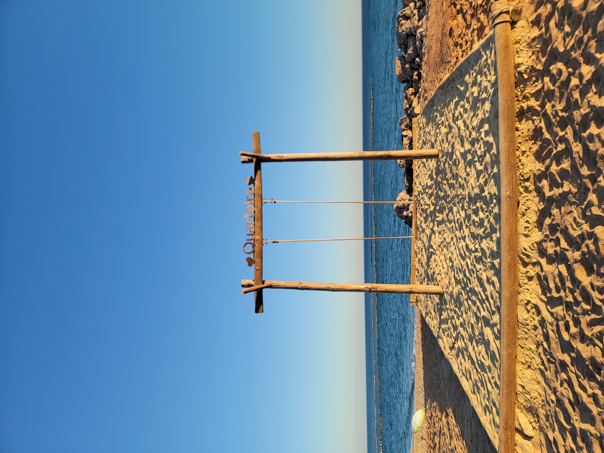 A swing set is centered on a sandy beach in Quarteira, Portugal, with the Atlantic Ocean and a clear blue sky in the background. The swing frame is made of rough wooden posts and beams, with two simple swings hanging from metal chains. A decorative sign with a heart and the word "Quarteira" is attached to the upper beam. The sand around the swing set is packed flat within a wooden border, while further out, the sand is textured with footprints and shadows cast by the low sun. Rocks are visible along the shoreline, and a series of buoys mark a boundary in the water. The water is calm and a lighter blue near the horizon, transitioning to a darker blue closer to the shore. The image captures a peaceful, late afternoon scene on a Portuguese beach.