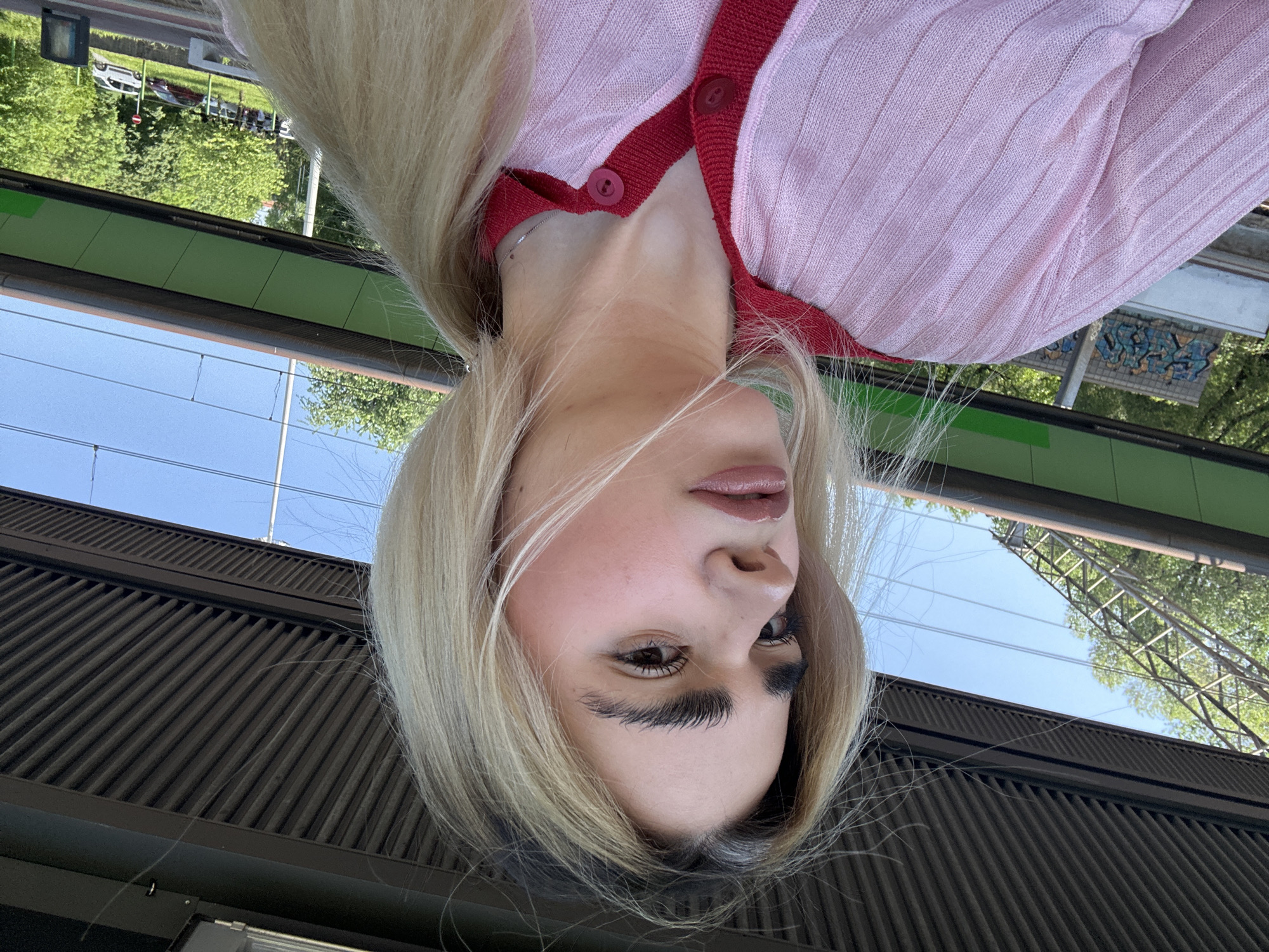 The image, correctly oriented, is a close-up, low-angle selfie of a young woman outdoors in Milan, Italy. She has long, straight blonde hair, dark, prominent eyebrows, and full lips with glossy, peachy-pink lipstick. She is wearing a light pink, finely ribbed cardigan with a contrasting red trim along the edges and red buttons, one of which is fastened. A thin silver chain is visible around her neck.
The background is layered, revealing an urban environment. In the immediate foreground, a dark brown, corrugated or slatted architectural surface is prominent, suggesting a wall or part of a structure she is near. Above this, a vibrant green horizontal panel extends across the mid-ground. A clear blue sky occupies the upper portion of the frame, crisscrossed by thin overhead power lines and featuring part of a large, metallic structural element or pylon on the right.
Much of the background scene is visible as a reflection, likely on a glass or highly polished surface in front of the woman. This reflection shows lush green trees, several parked cars, a light-colored building, and a dark trash can, indicating an outdoor, public setting such as a street or platform area. Patches of graffiti with blue and yellow lettering are faintly discernible on a distant wall in the reflected scene.
The bright, clear light suggests it is daytime with sunny weather. The combination of architectural features, power lines, and reflections of vehicles points to a setting near a public transportation hub, such as a train or tram station, consistent with the provided location context of Milan, Italy.