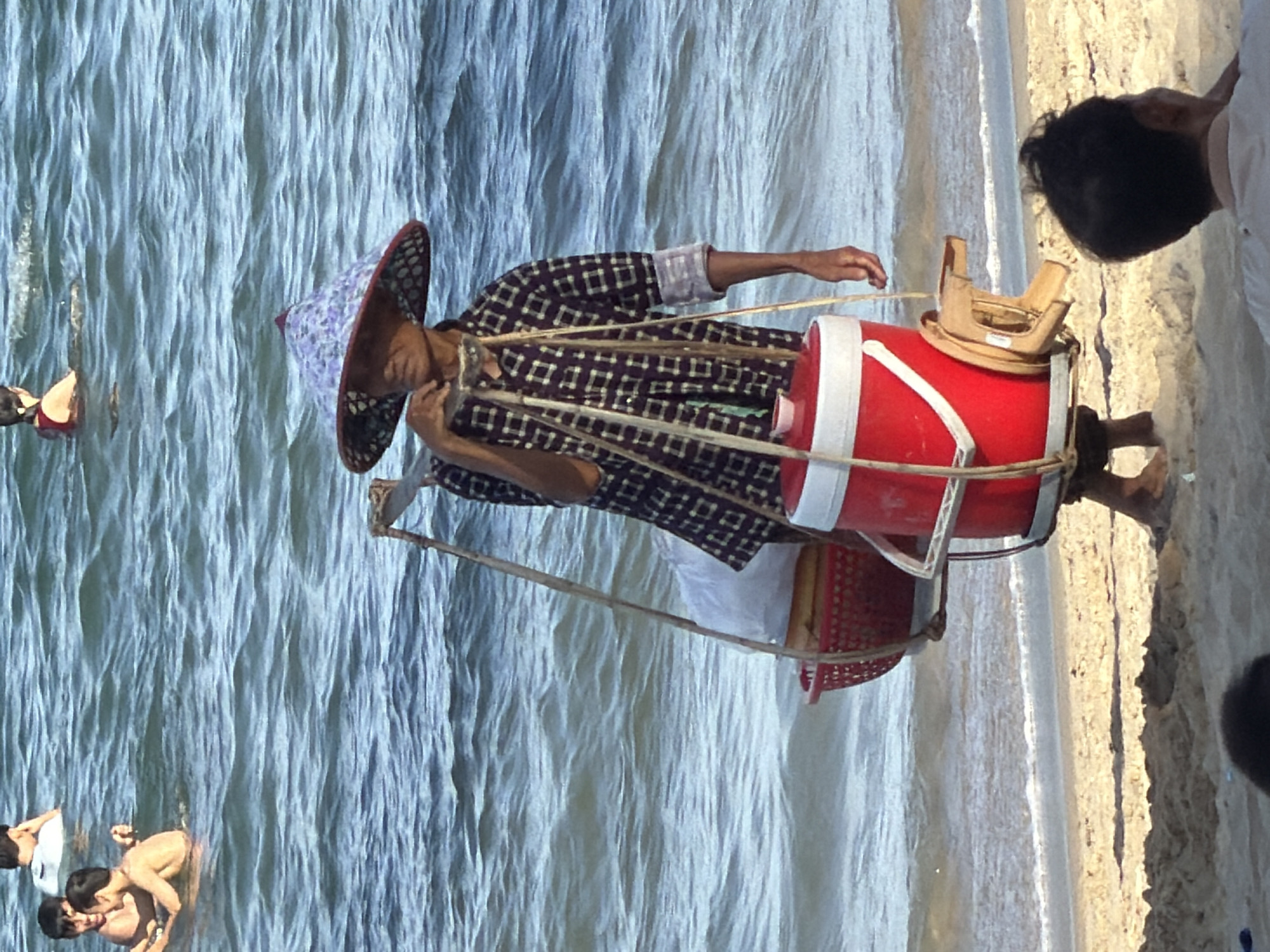 A person wearing a conical hat and a plaid shirt walks along a sandy beach next to the ocean. The person is carrying a yoke over their shoulders with two baskets hanging from either end. The left basket is red and contains white bags. The right basket is a red cooler with a white lid, and a small wooden stool is attached to its side. Several people are visible in the water in the background. The scene appears to be in Nha Trang, Vietnam, based on the presence of conical hats and the general beach environment.