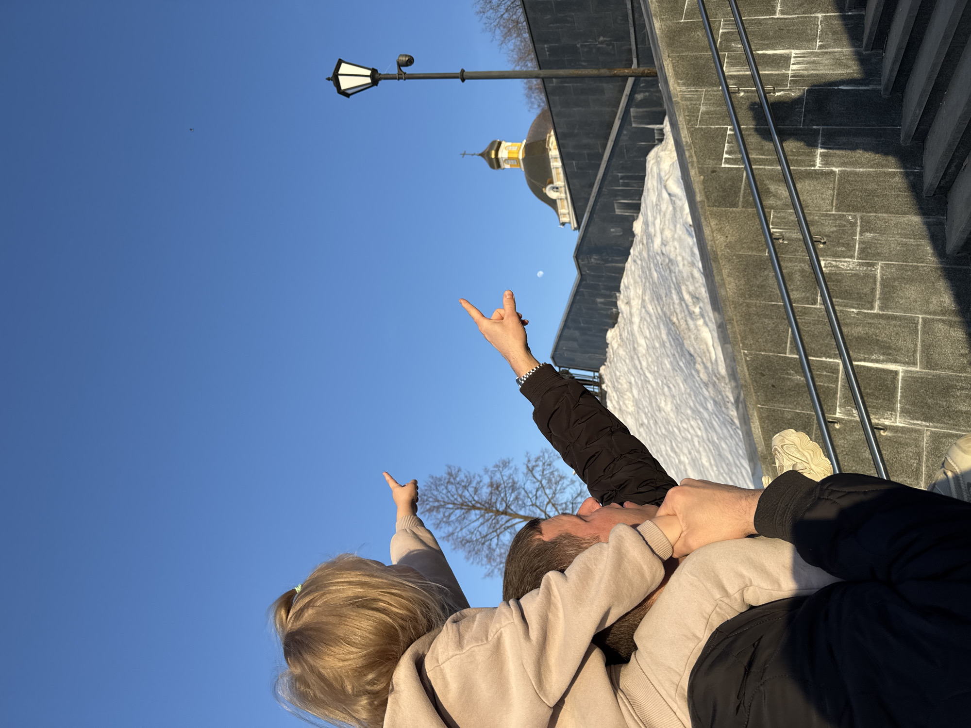 A ground-level, medium-shot, tilted photo taken in Perm, Russia, focuses on two people from the chest up, embracing and gazing upward. The person on the left, with blonde hair pulled back, wears a light brown hoodie and points a finger towards the sky. To their right, the other person, with short brown hair, is dressed in a dark jacket and raises their hand in a "rock on" gesture.
In the background, a clear blue sky dominates, with a streetlamp and the dome of a church visible above a gray stone wall covered with snow. The gray stone wall includes metal railing.