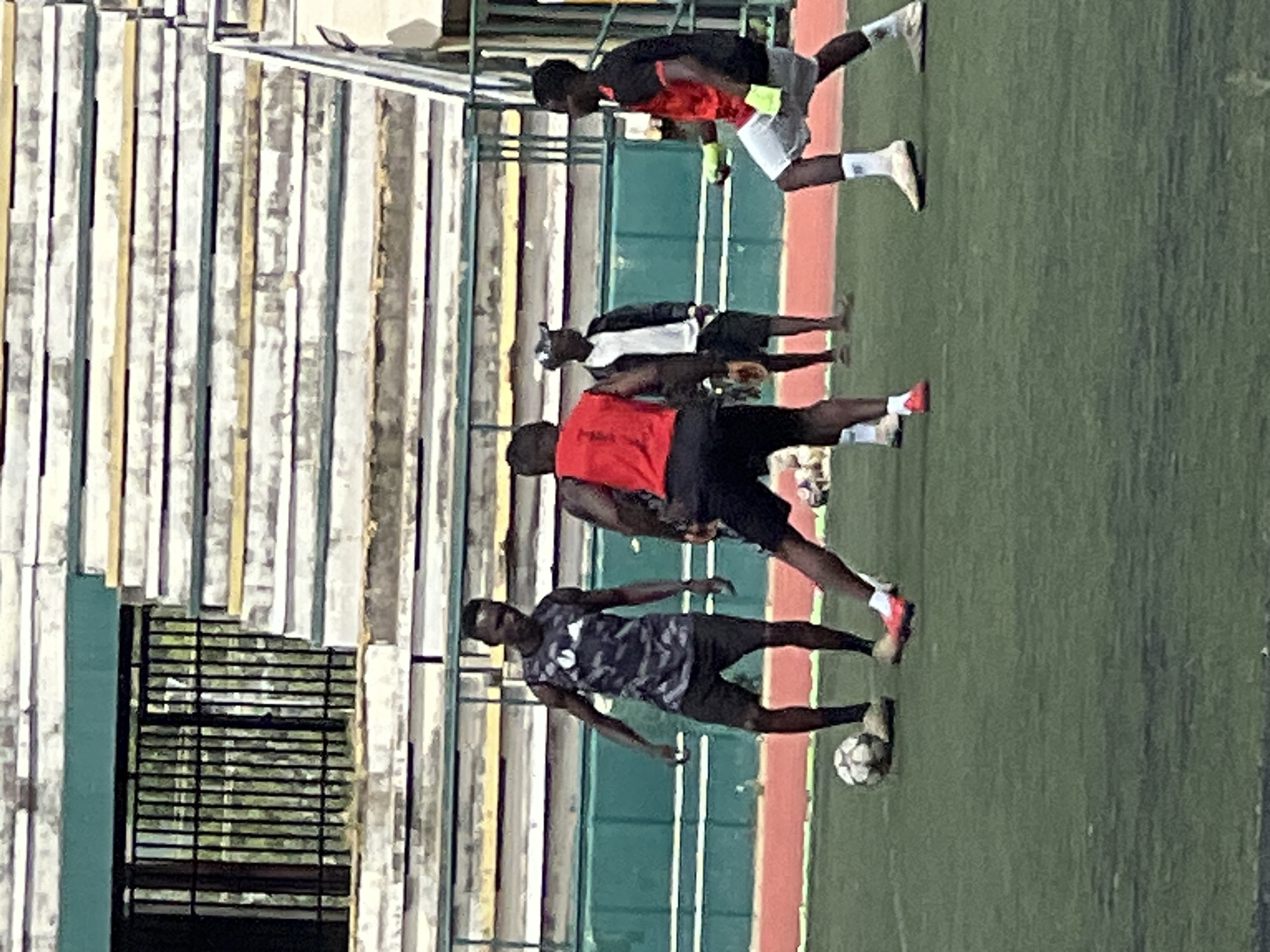 Four individuals are on a green turf field, possibly a sports ground, at the New Stadium in Nigeria. The field is marked with a red border. In the background, there are tiered stadium seats, some of which appear to be under construction or in disrepair, with visible metal bars and scaffolding.
The individuals are engaged in what appears to be a soccer practice. One person on the left is wearing a dark camouflage sleeveless shirt and black shorts, standing with a soccer ball at his feet. To his right, a person in a red sleeveless jersey and black shorts is captured mid-stride, facing away from the camera. Further right, another individual in a white long-sleeved shirt and black shorts with a cap is standing, observing. On the far right, a person in a red and black jersey with white shorts is facing towards the left of the frame, holding what appear to be bright yellow goalkeeping gloves. Another soccer ball is visible near this individual. The lighting suggests it is daytime, likely overcast.