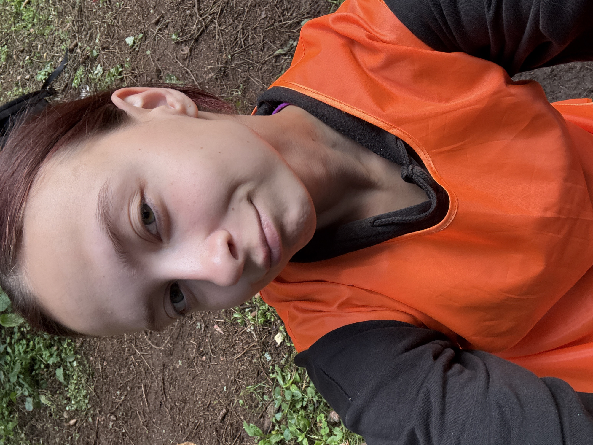 The image shows a young person wearing a black long-sleeved shirt and an orange vest. They are lying down outdoors, with their head tilted towards the camera. The background consists of dirt and some greenery, suggesting an outdoor setting like a park or forest. The person is looking at the camera with a slight smile. The lighting suggests it is daytime, possibly overcast. There is no visible text in the image. The overall mood appears relaxed and casual. The person's hair is a reddish-brown color and is pulled back from their face. The orange vest is bright and stands out against the darker clothing and natural background.