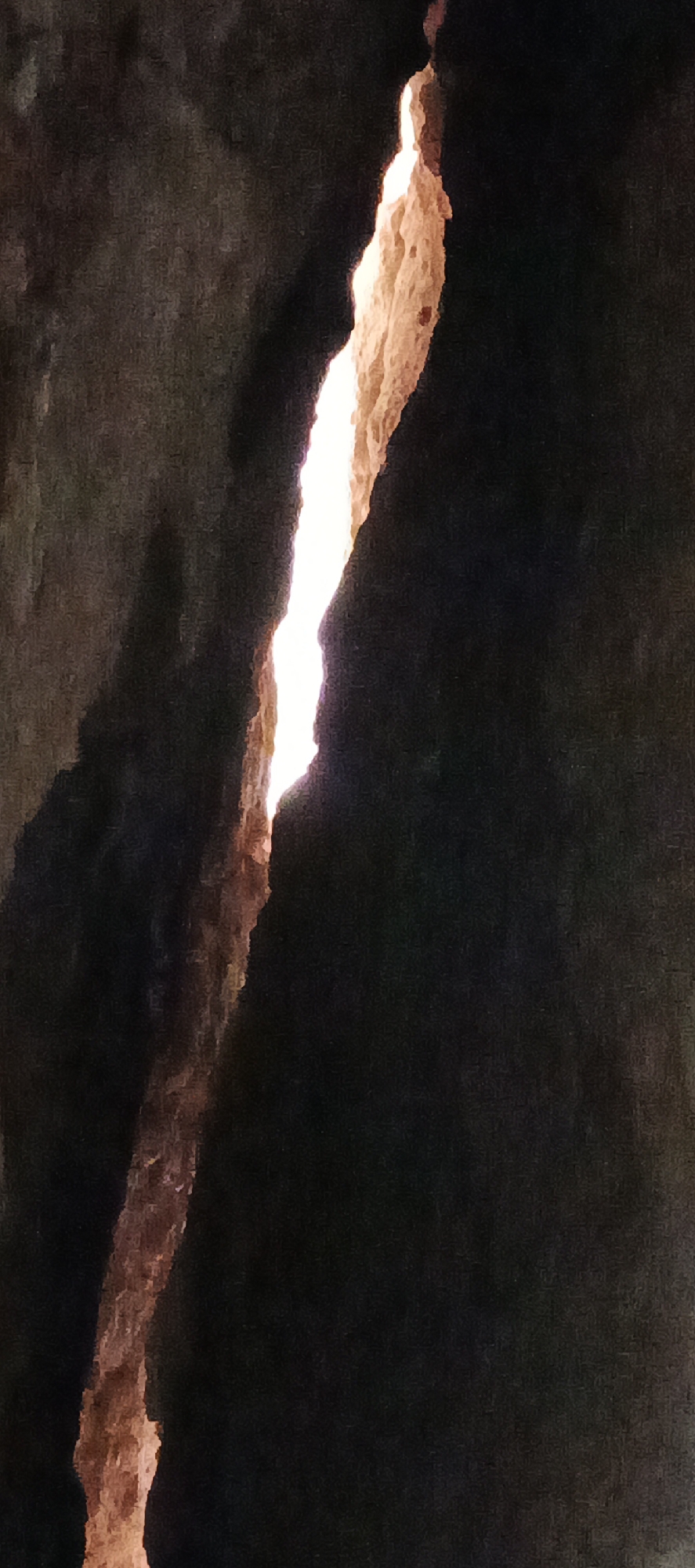 The image depicts a striking natural formation, likely a deep crevice or crack in rock or earth. A narrow, vertical fissure runs through the center, glowing intensely with a warm, bright light. The light illuminates the rough, textured edges of the crack, which appear in shades of sandy brown and orange. The surrounding area is enveloped in deep, dark shadows, creating a stark contrast and emphasizing the luminosity of the opening. The overall setting suggests a dark, enclosed space, such as a cave or canyon, with an unseen external light source, possibly sunlight, penetrating through the narrow gap. There are no visible people, objects, or text. The scene captures the dramatic effect of light piercing through darkness, highlighting the natural textures of the rock.