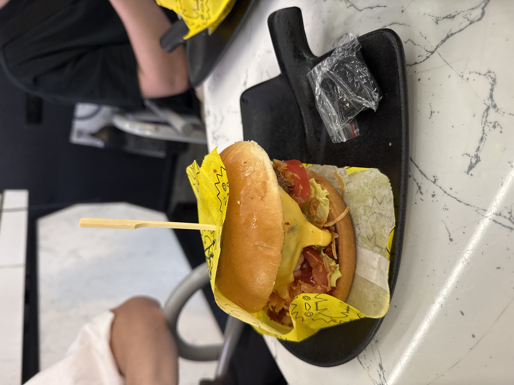The image depicts a close-up of a gourmet hamburger served on a black, shovel-shaped plate. The hamburger is partially wrapped in yellow paper with a cartoon monster print. It features a golden-brown bun, melted cheese, lettuce, tomato, and other burger ingredients. A wooden skewer pierces the top bun. Two small, sealed plastic bags containing dark granular material are placed on the plate to the right of the hamburger. The plate rests on a white marble-patterned table. In the background, out of focus, are two individuals seated at a table. One person is wearing a white shirt, and the other is wearing a black t-shirt. The overall setting suggests a restaurant environment in Saint Petersburg, Russia.