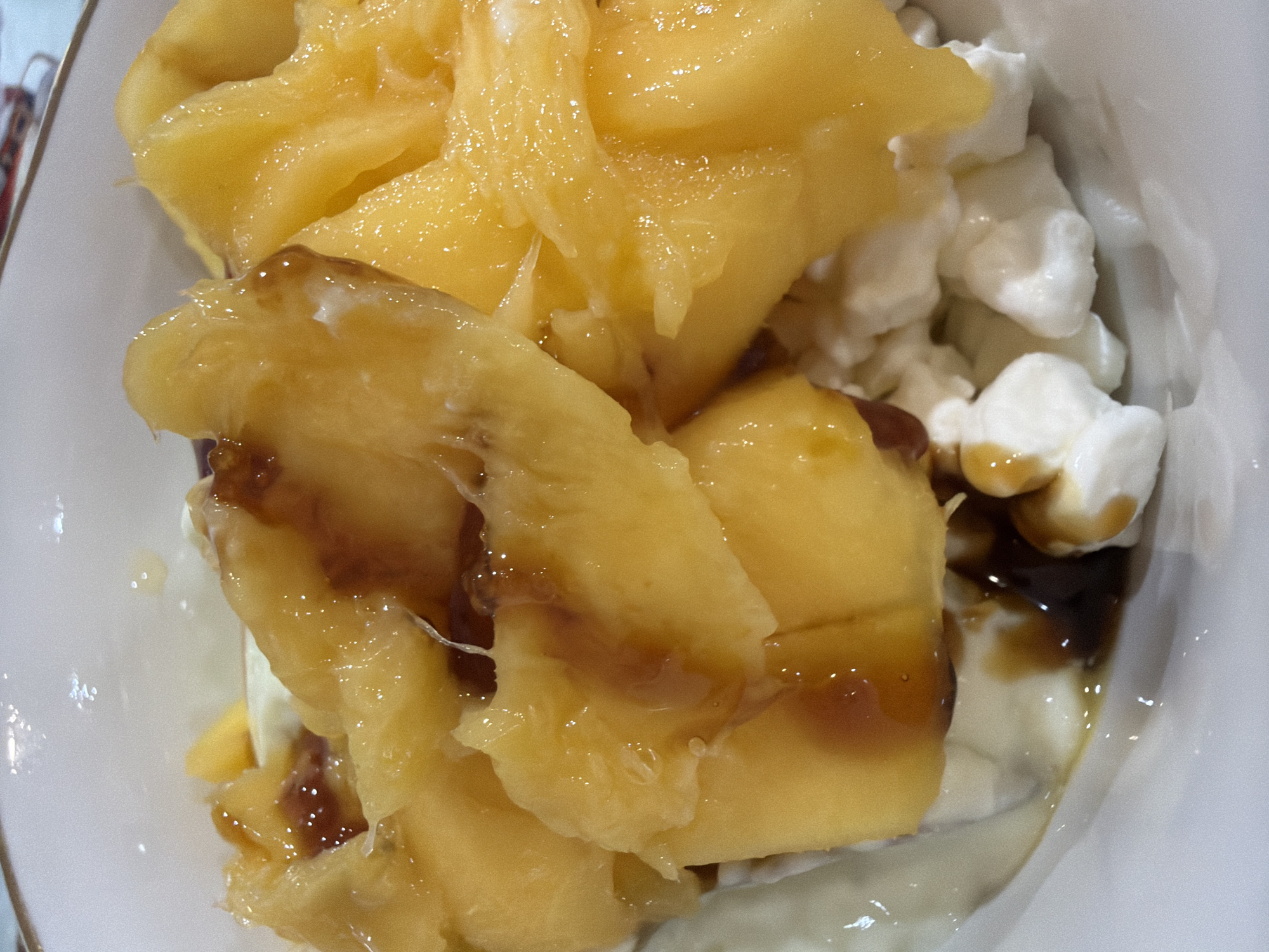 A close-up overhead shot reveals a white bowl filled with a dish consisting of cottage cheese, sliced yellow peaches, and a drizzle of dark brown syrup. The peaches are cut into segments and layered on top of the cottage cheese. The syrup is drizzled unevenly, coating parts of the peaches and pooling in the spaces between the cottage cheese curds. The bowl appears to be ceramic, judging by its smooth, glossy surface. The image captures a simple food presentation, likely prepared and photographed indoors. There is no indication of a location in Chaikovsky, Russia.