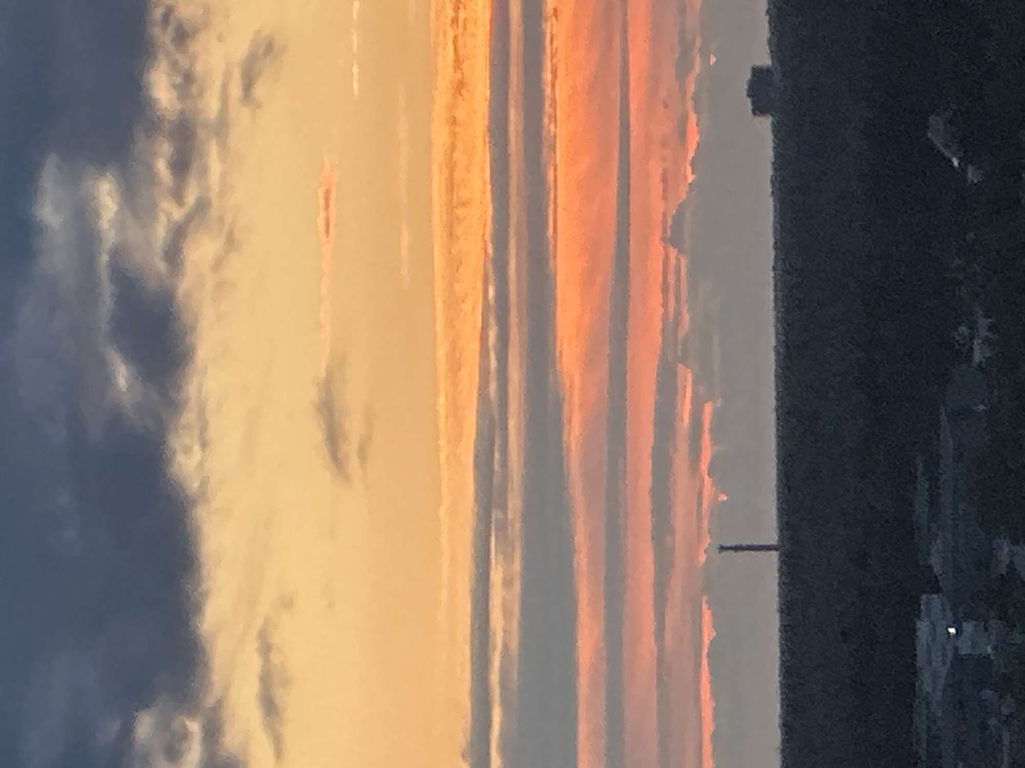 The image depicts a sunset over Podolsk, Russia, characterized by a dramatic sky filled with layers of clouds illuminated by the setting sun. The uppermost portion of the sky is a deep, brooding blue, transitioning to vibrant shades of orange and pink as it descends. Horizontal bands of clouds create a striped effect across the sky, with some tinged with fiery red and others a soft grey. In the distance, silhouetted against the colorful sky, is a dark, treed landscape. A tall, slender tower, likely a radio or transmission tower, rises from the treeline on the left side of the frame. To the right of the tower, a low-profile, dark building is visible on the horizon. The foreground shows the dark expanse of the treeline, with hints of urban development, including buildings and lights, at the very bottom of the image. The overall scene is one of natural beauty, with the dramatic sky dominating the composition.