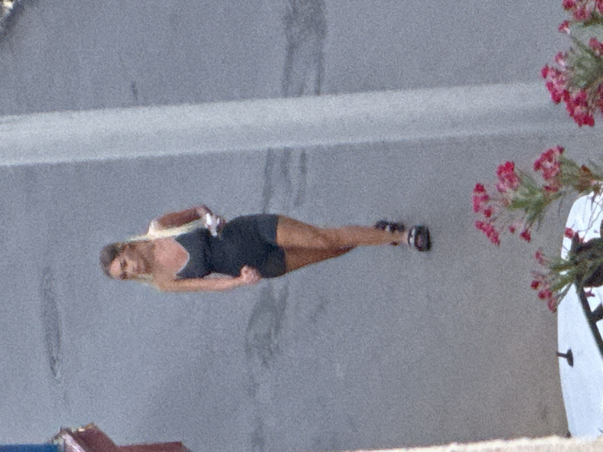 A blurry, medium-angle shot shows a woman with blonde hair wearing a dark one-piece athletic outfit and sandals, jogging on a gray asphalt surface. She is holding a white object in her right hand. The background is out of focus, with a hint of a sidewalk and some pink flowers in the foreground. The scene appears to be in a city, possibly Budva, Montenegro, given the presence of buildings and a street.