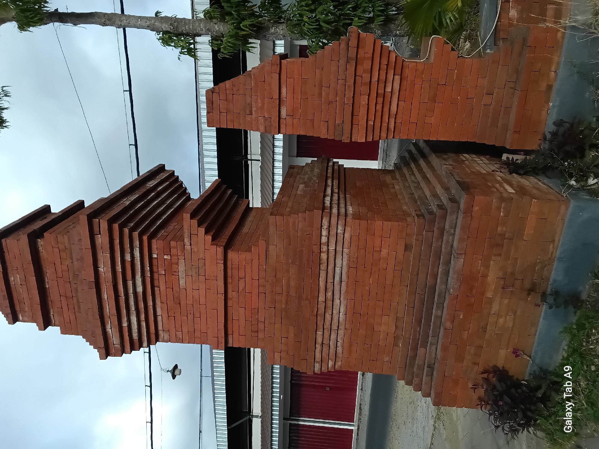 In Bendorejo, Indonesia, a brick gate structure stands prominently, featuring tiered layers of reddish-brown bricks with some lighter-colored stones interspersed. The structure appears to be an entryway, with one side notably broken and angled away from the main body, revealing a dark, recessed opening. To the left of the gate, a street and dark red, roll-up doors of a building are visible. A tree with a slender trunk and green foliage rises to the right of the gate, silhouetted against a cloudy sky. A single bulb hangs from a wire above and to the left of the gate. The foreground shows patches of grass and dark green, leafy plants. The "Galaxy Tab A9" watermark is present in the bottom left corner.