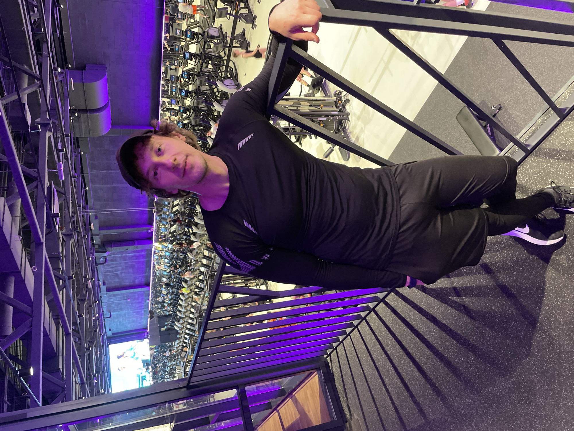 A young man with curly blonde hair, wearing a black long-sleeved shirt, black shorts, and a black baseball cap, leans against a black metal railing in a gym. The man is positioned in the foreground, with his right arm resting on the railing and his left hand partially out of view. The background is a large gym with rows of cardio machines, including treadmills and ellipticals, illuminated by purple lighting. A large screen displaying an image of people is visible in the far background. The gym floor is dark and speckled, with distinct shadows cast by the railing.