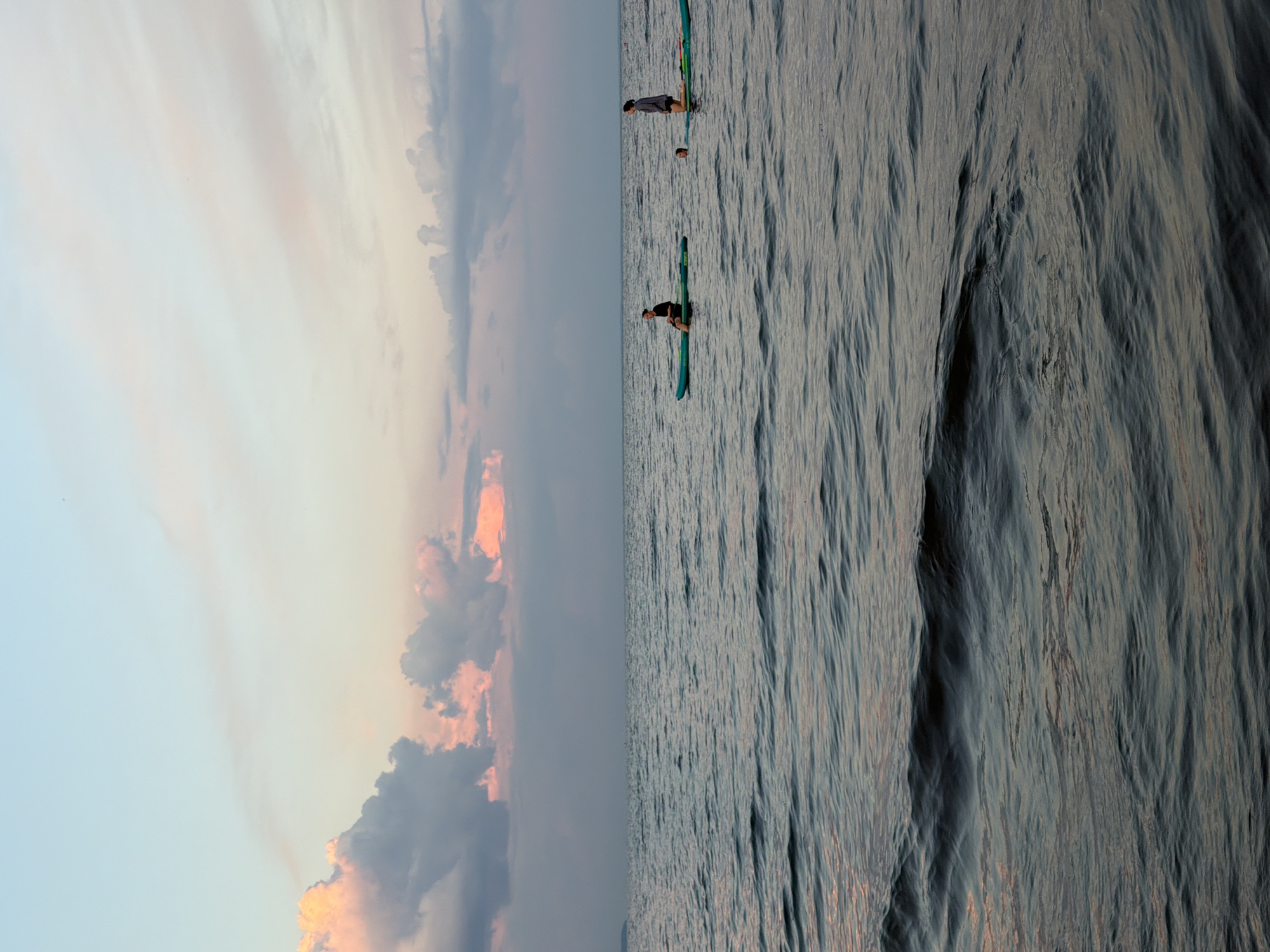 Two individuals are paddleboarding on a calm body of water. The person on the left is sitting on their paddleboard, facing to the right. The person on the right is kneeling on their paddleboard, facing forward. The sky above is a pastel gradient of blue and pink, with some fluffy clouds in the distance. The water is dark and reflects the colors of the sky. The scene appears to be at sunset or sunrise. This image was taken in Laishevo, Russia.