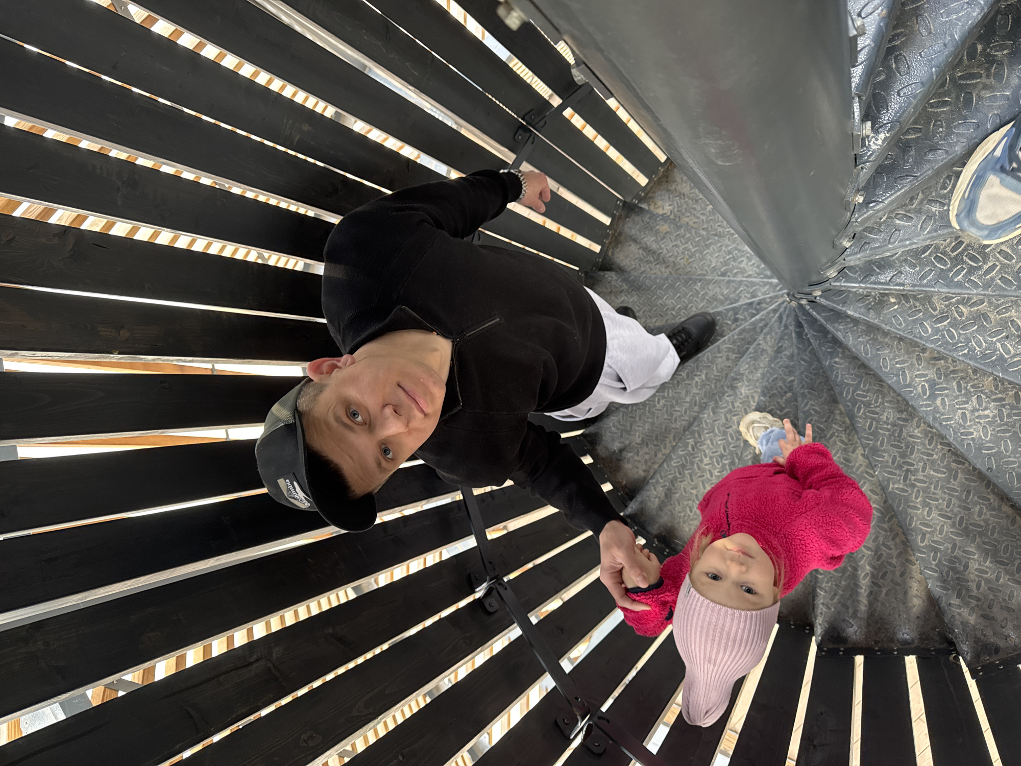 A man and a young child are standing on a spiral staircase inside a building in Perm, Russia. The man is wearing a black jacket, grey pants, and a baseball cap. The child is wearing a pink hat and a red jacket. The man is holding the child's hand. The staircase is made of metal and wood.