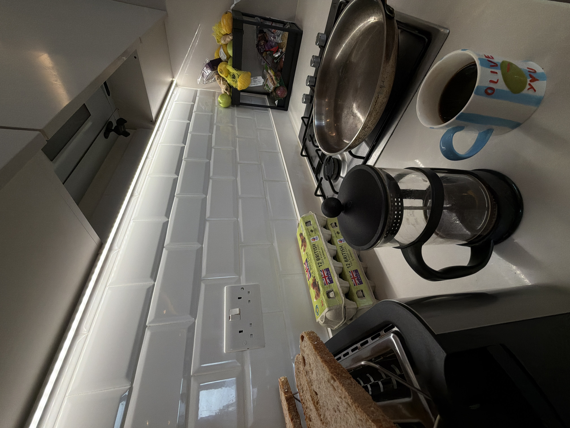 The image shows a kitchen countertop scene. In the background, there are white subway tiles and kitchen cabinets. Fruits are stored in a black wire basket. On the countertop is a gas stove with a frying pan on it. A mug with blue stripes and the words "OLIVE YOU" sits near the pan. A French press coffee maker is also present. A carton of "12 British Free Range Eggs" is next to the coffee maker. At the bottom of the image, there is a toaster with slices of toast in it. A UK-style electrical outlet is visible on the tiled wall. There is no indication that this scene is in London.