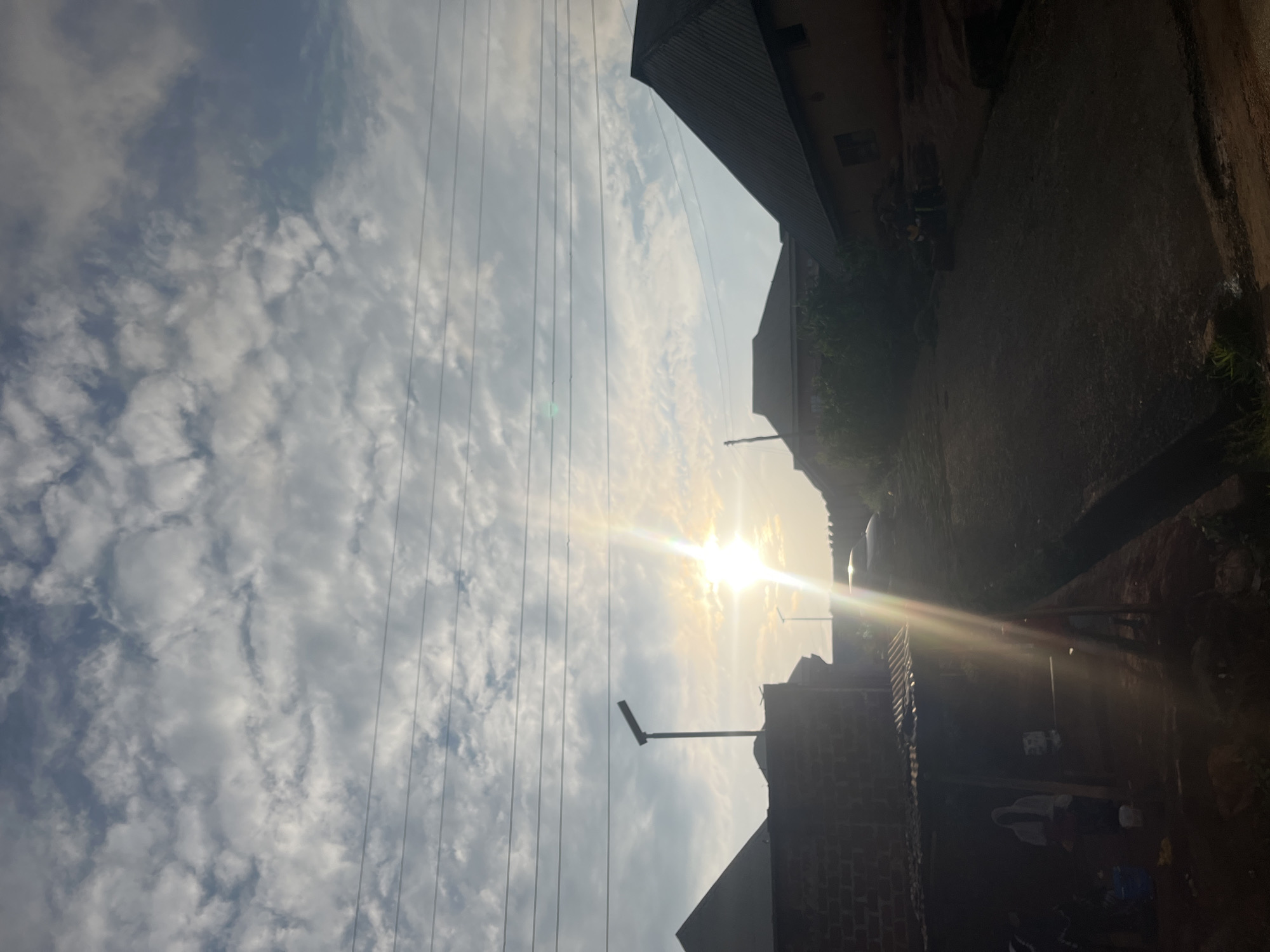 This image captures a street scene in Gembu, Nigeria, bathed in the warm glow of the sun. The sun is positioned high in the sky, casting bright rays that create a lens flare effect. The sky is filled with fluffy, white clouds against a light blue backdrop, suggesting fair weather.
The foreground and right side of the image are dominated by the shadows and shapes of buildings, with roofs and walls visible. Some of these structures appear to be made of brick, while others have metal roofing. A few sparse green plants are visible near the base of some buildings, indicating some vegetation despite the urban setting.
Several power lines are strung horizontally across the image, a common sight in many urban and semi-urban environments. A street lamp is also visible, its arm extending outwards. The overall impression is that of a residential or mixed-use area.
The low angle of the sun and the long shadows suggest it is either early morning or late afternoon. There are no people clearly visible in the image, but the presence of the buildings and power lines implies human habitation and infrastructure. The scene evokes a sense of calm and a typical day in a Nigerian town. No visible text is present.