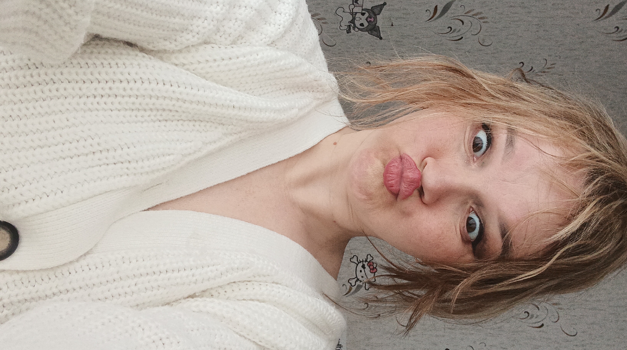 An indoor close-up selfie depicts an adult individual with light blonde hair, appearing disheveled, and fair skin, located in Ольховка, Russia. The person is wearing a white, chunky knit cardigan with a V-neck. Their facial expression includes wide, slightly cross-eyed eyes with dark eyeliner, and lips pursed into a "duck face." The background consists of a light grey wall covered with wallpaper featuring a subtle dark grey, swirling floral or vine pattern. Two distinct cartoon character decals are visible on the wallpaper: one resembling Kuromi holding a cup is positioned on the left, and another resembling a pirate-themed Hello Kitty with an eye patch is visible on the right, partially obscured by the individual's hair.