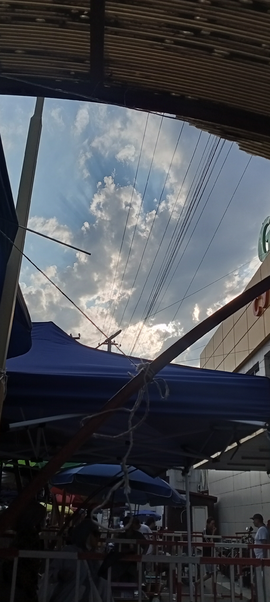 The image captures a street scene in Sokuluk, Kyrgyzstan, beneath a corrugated metal overhang. In the foreground, a blue tarp covers market stalls, supported by a wooden pole tied with rope. Beyond this, a building with a beige facade and a large circular green logo is visible. The sky is partly cloudy, with sunlight filtering through, illuminating several power lines crisscrossing the frame. In the background, people are seen gathered at what appears to be a market, with various stalls and umbrellas providing shade. The overall atmosphere suggests a busy commercial area.