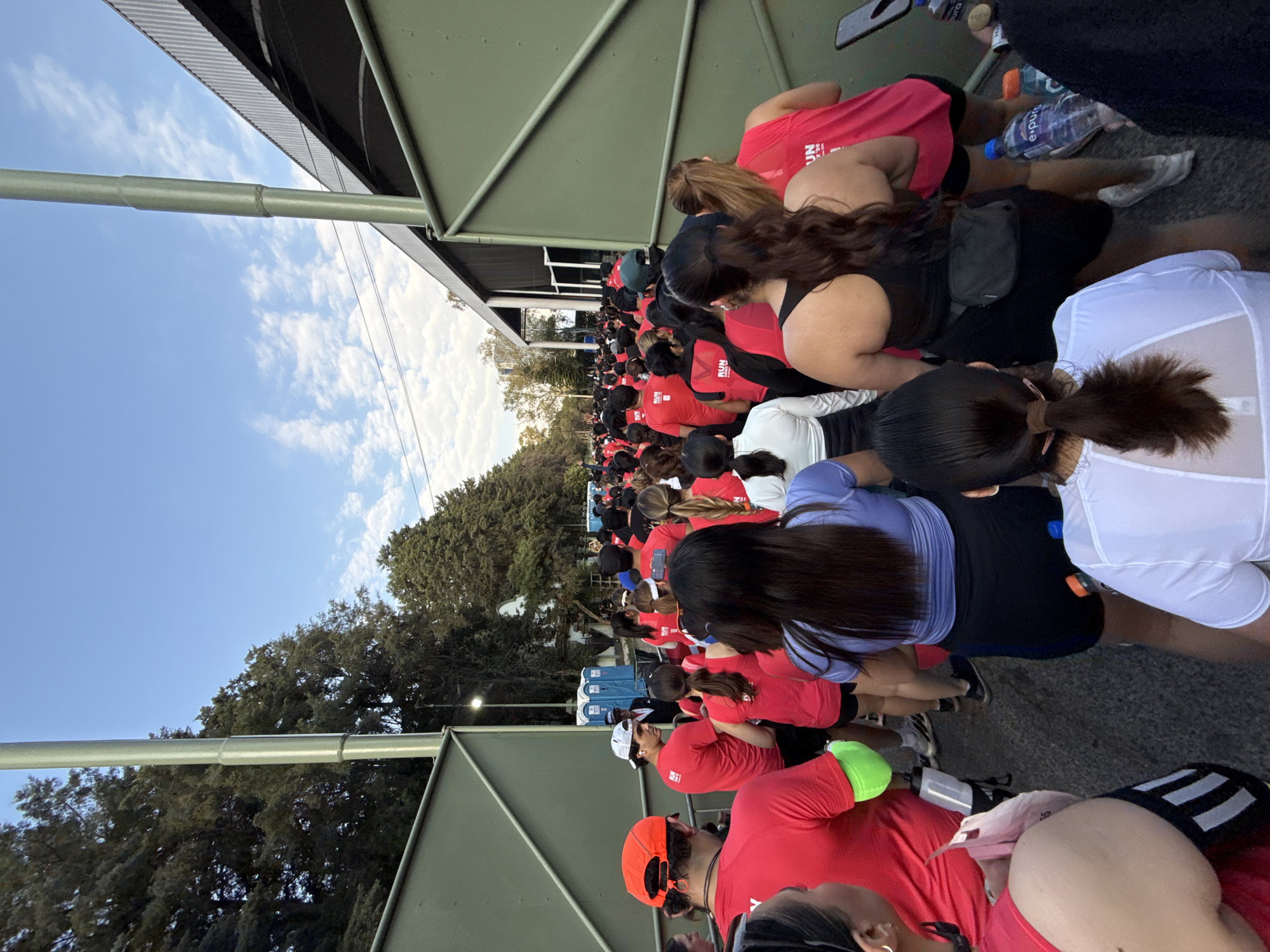A dense crowd of people, many wearing red shirts, are gathered outdoors in Mexico City, Mexico. They appear to be lining up or waiting. Some are sitting. Some are holding water bottles. The setting includes trees, a blue portable toilet, a building structure with metal supports, and a sky with blue patches and scattered clouds.