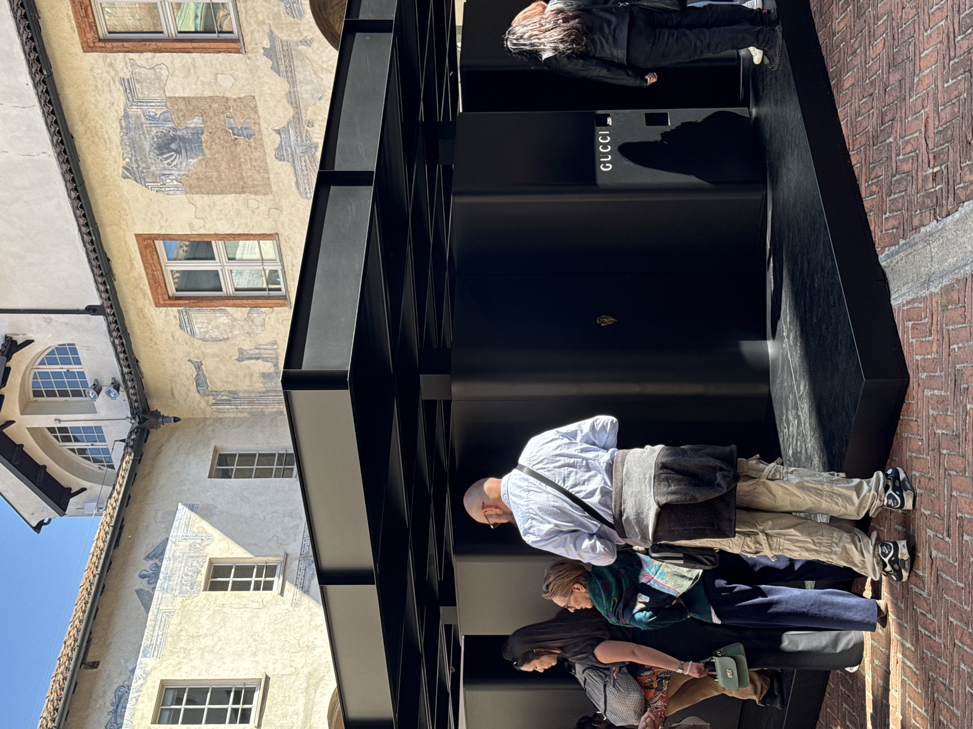 The image captures a scene outside a Gucci store in Milan, Italy. The store's facade is modern and black, with the "GUCCI" logo prominently displayed. People are standing in front of the store, likely window shopping or waiting to enter. The architecture in the background features older buildings with traditional Italian designs. The street is paved with bricks. The sky is blue and sunny.